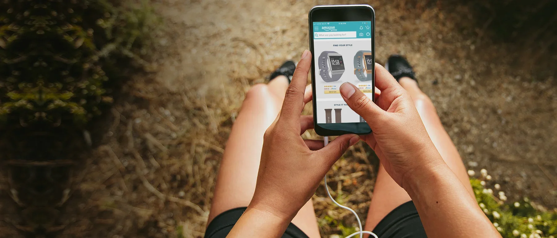 A person sitting outdoors on a dirt path, shopping on their phone for smartwatches on Amazon, with legs and feet visible, wearing shorts and sneakers, and holding a phone connected to earphones.