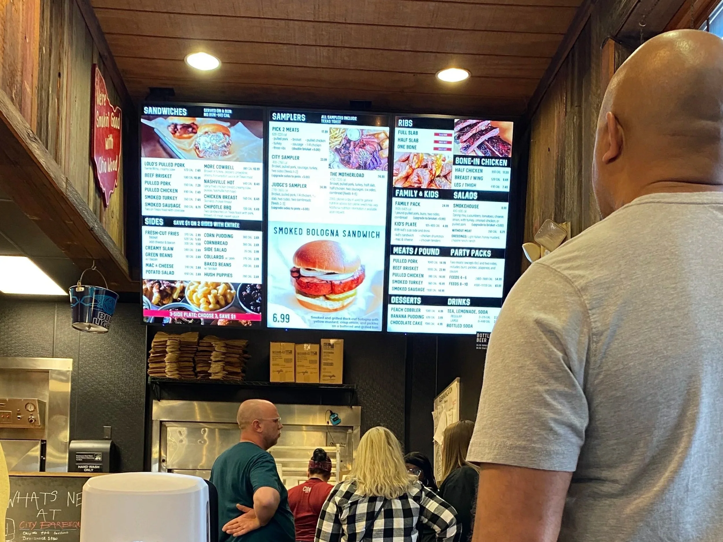Digital menu board at a barbecue restaurant with various meat, sides, and drink options, visible in a restaurant setting with customers waiting in line.