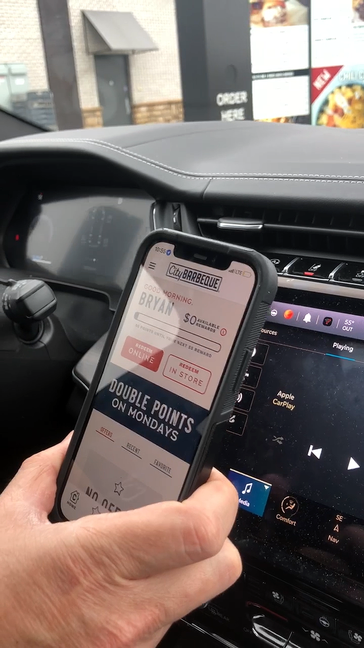 Inside a car, a person's hand holds a smartphone with a rewards app open, showing a 'City Barbeque' loyalty program and offers. In the background, the car's dashboard and an exterior fast food restaurant can be seen through the windshield.