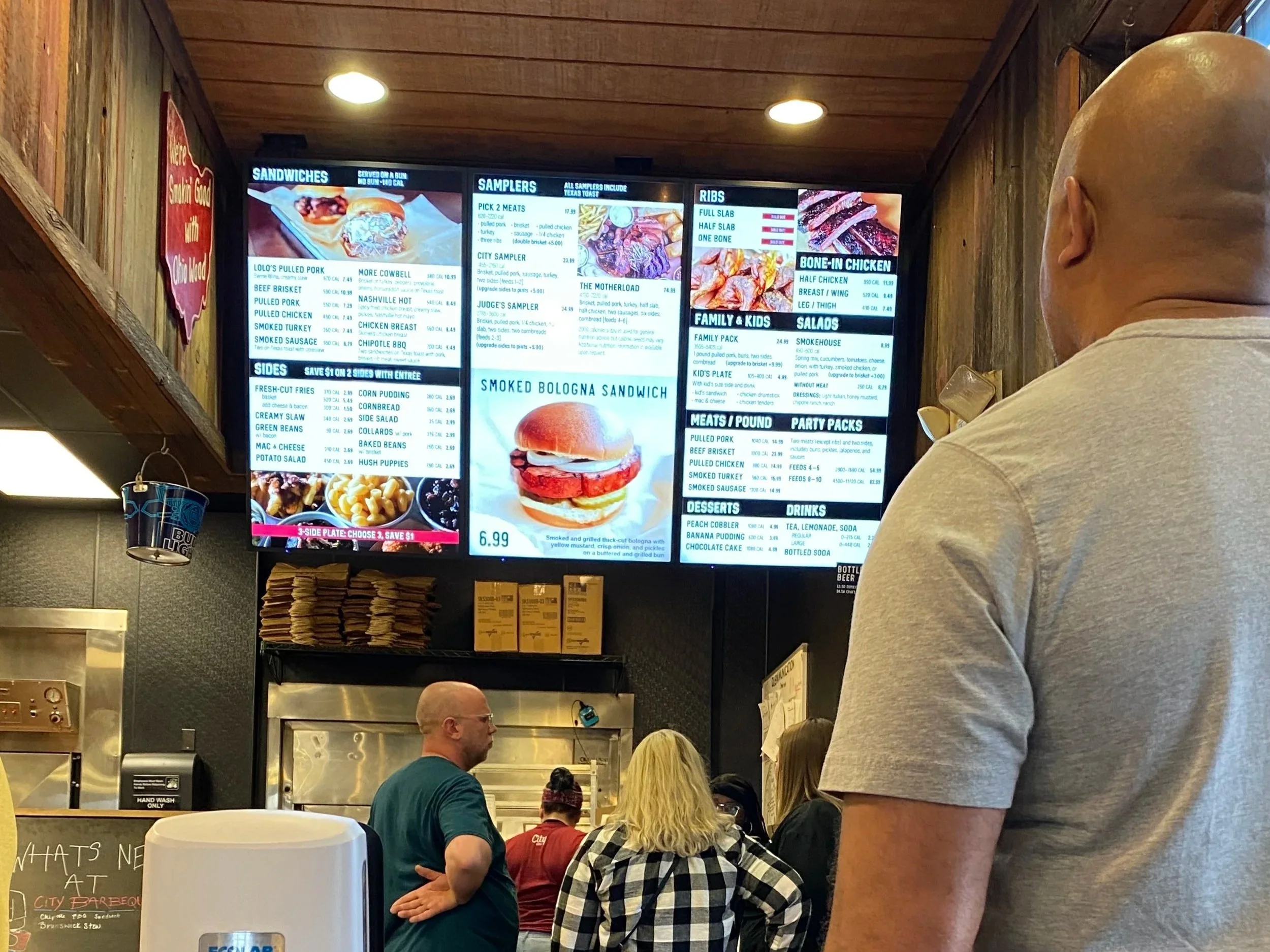 A digital menu board displaying various food options in a restaurant, with people standing and ordering in the foreground and background.