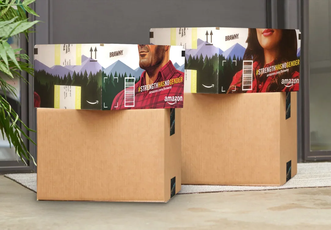 Two cardboard boxes with Amazon shipping labels and mountain landscape artwork on the flaps, placed on a light-colored floor near a plant and a gray wall.