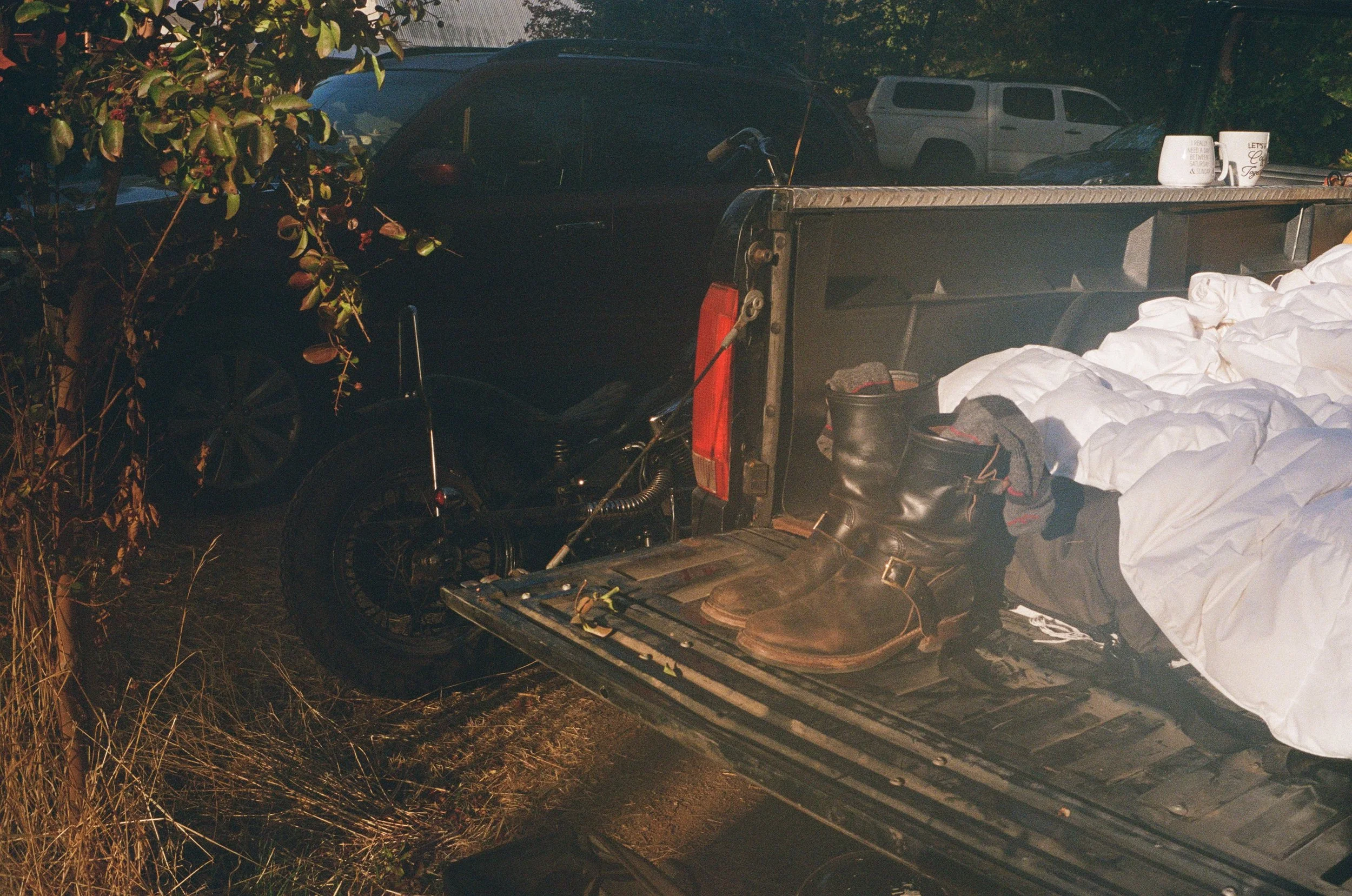 A black pickup truck parked outdoors with items in its bed, including white bags or blankets, and a green container. Several other vehicles are visible in the background, and there are trees and bushes nearby. The scene appears to be during the daytime or early evening.
