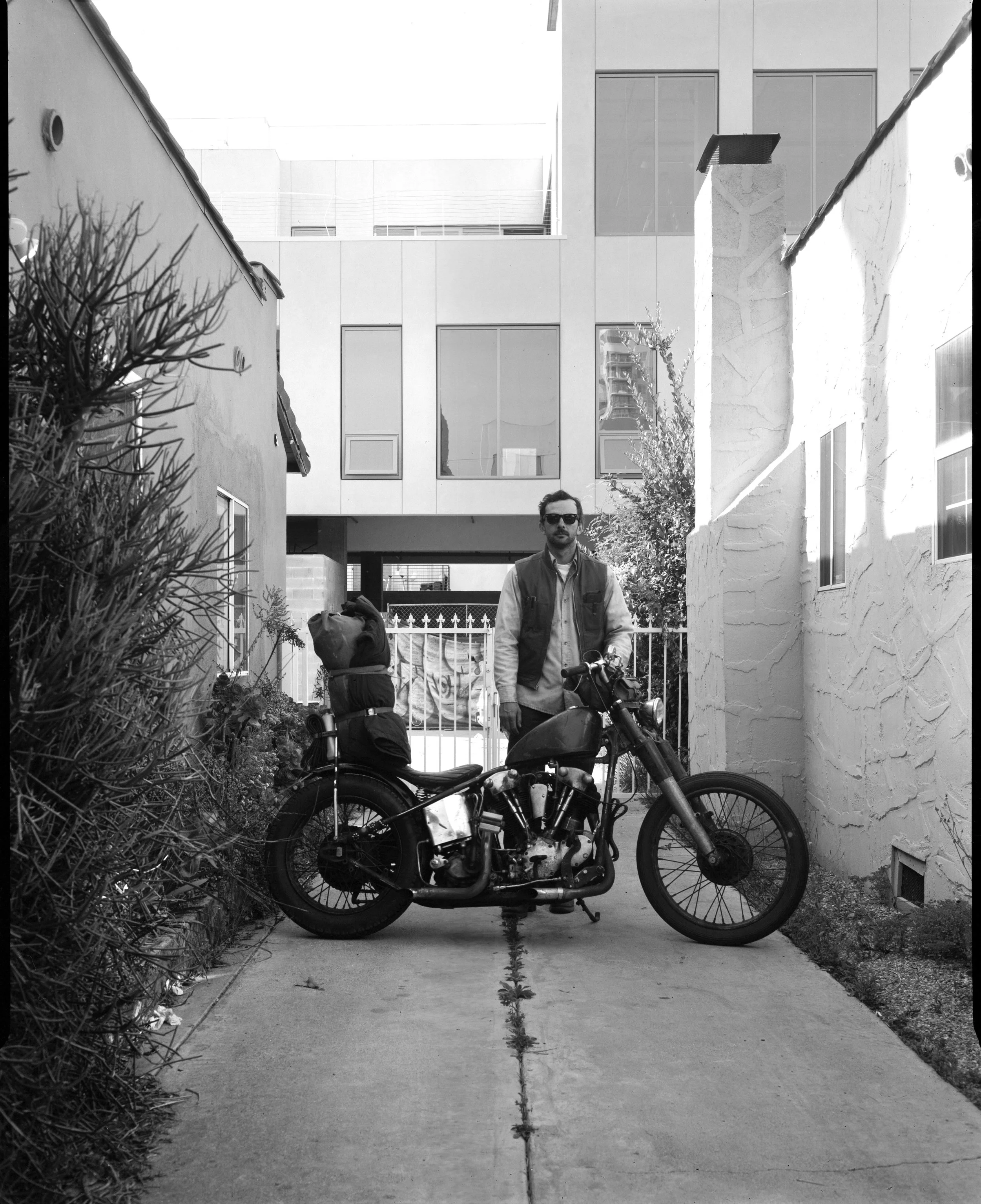 A man with sunglasses and a vest standing next to a motorcycle on a sidewalk, with residential buildings in the background, a garden, and a gate.