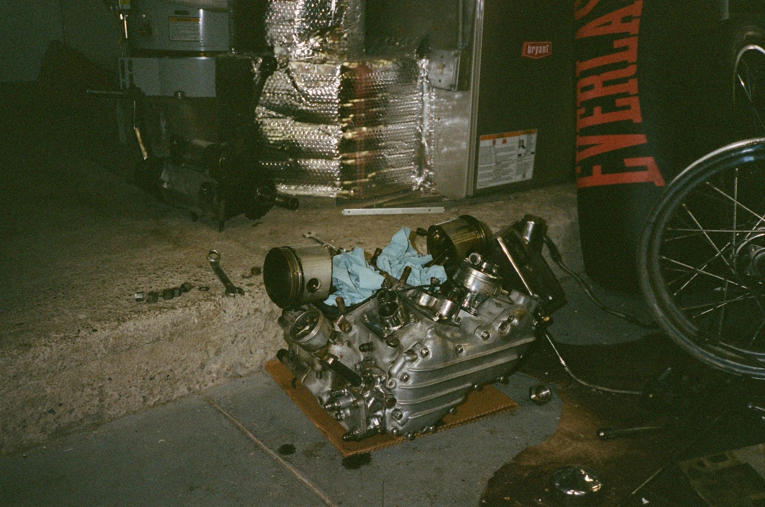 A disassembled engine placed on a cardboard on the floor, with various engine parts scattered around. In the background, there's a metallic box and a laundry basket with the words "Laundary" printed on it.