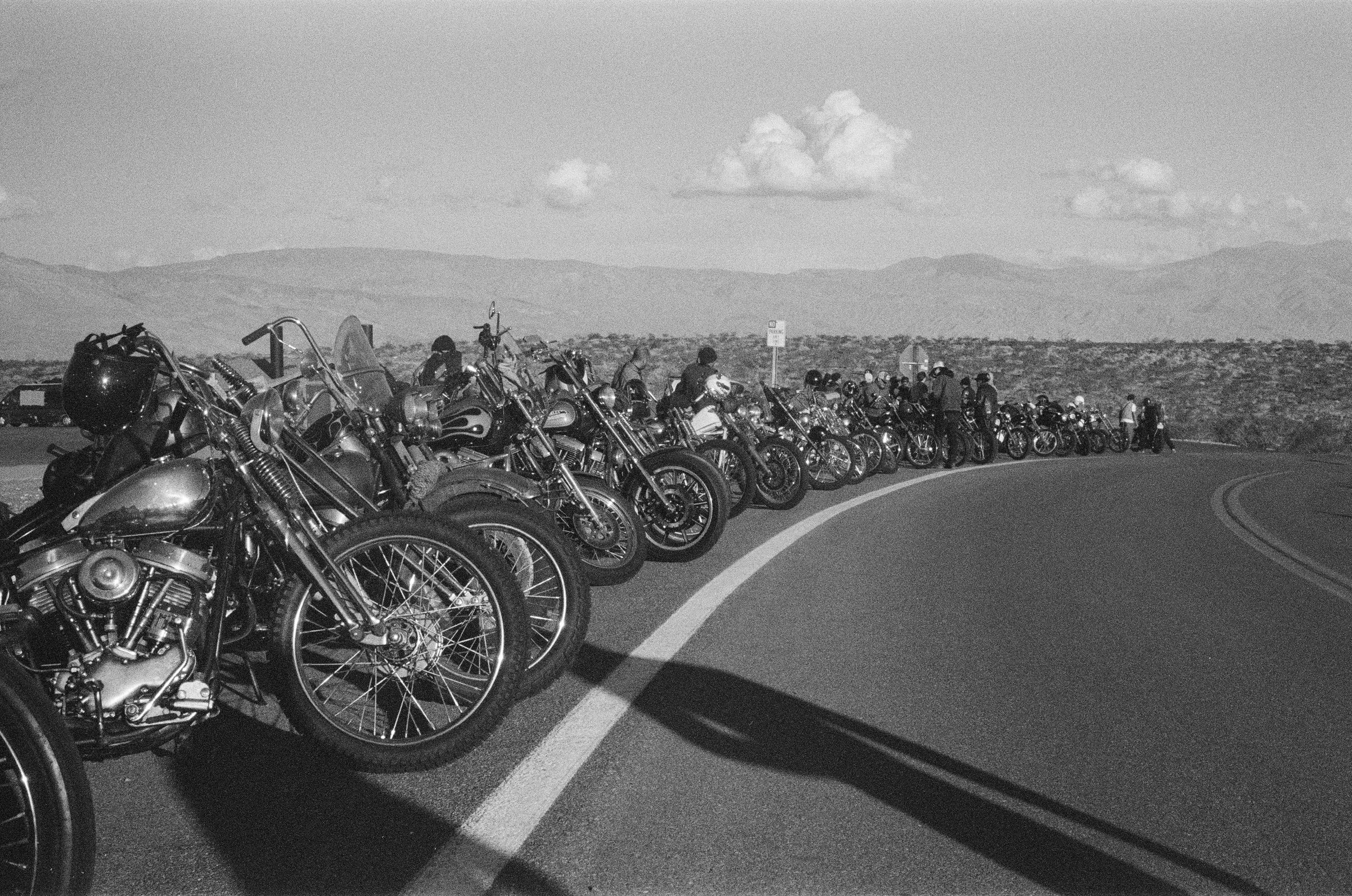Line of motorcycles parked along a curved road with mountains in the background and clouds in the sky.