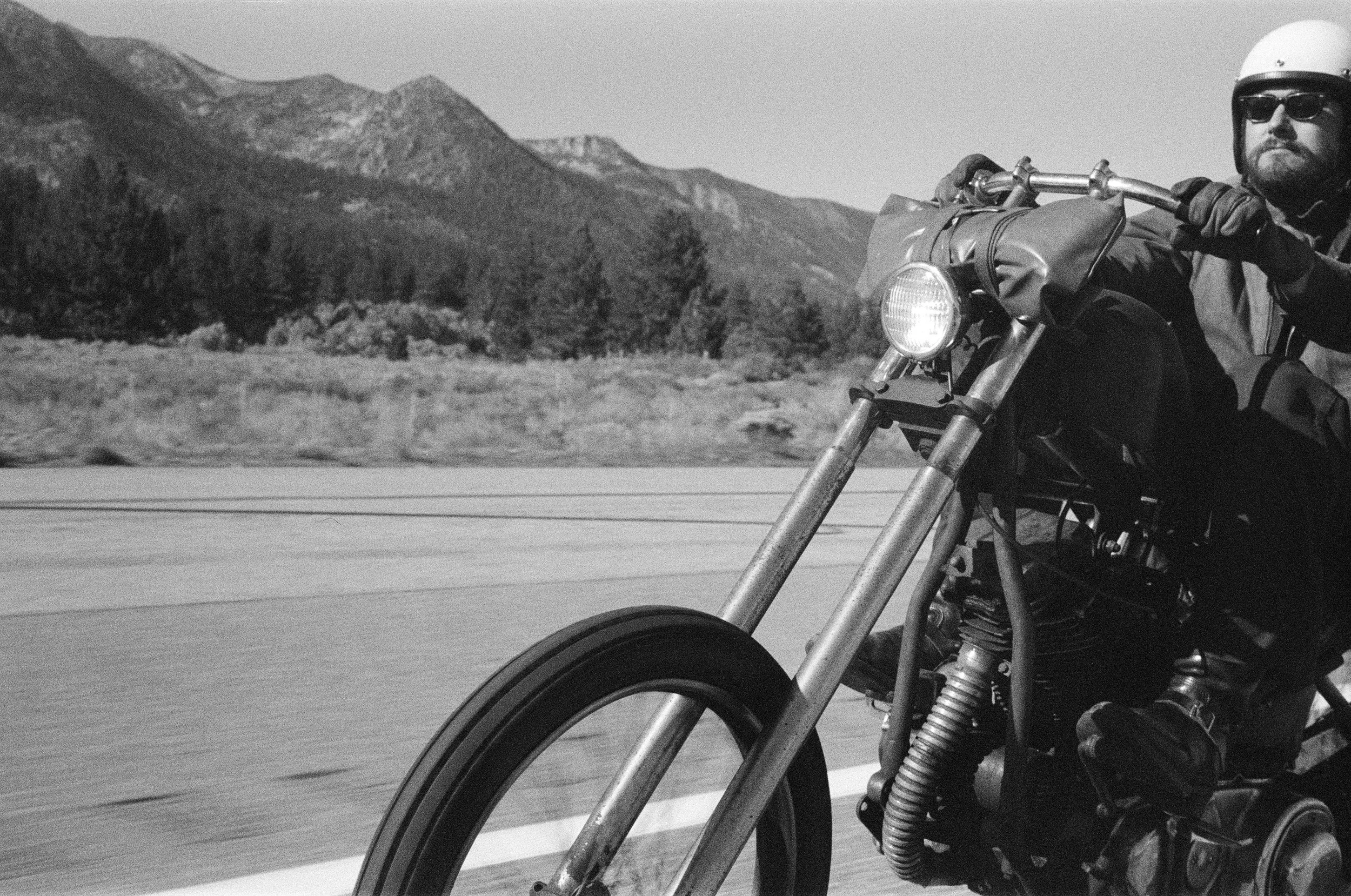 A man wearing sunglasses, a helmet, and gloves riding a motorcycle on a mountain road with mountains and trees in the background.