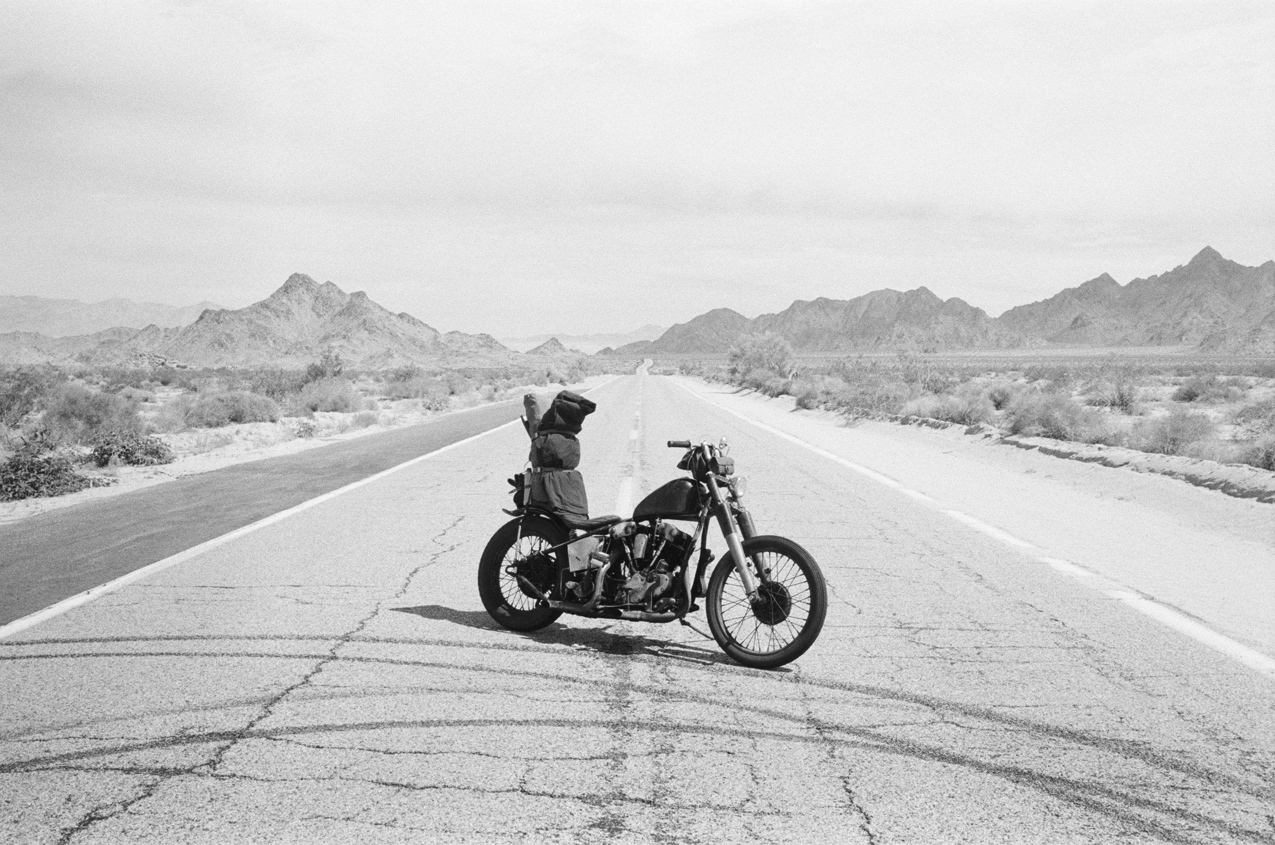 A motorcycle with gear and a backpack parked on an empty desert road with mountains in the background, in black and white.