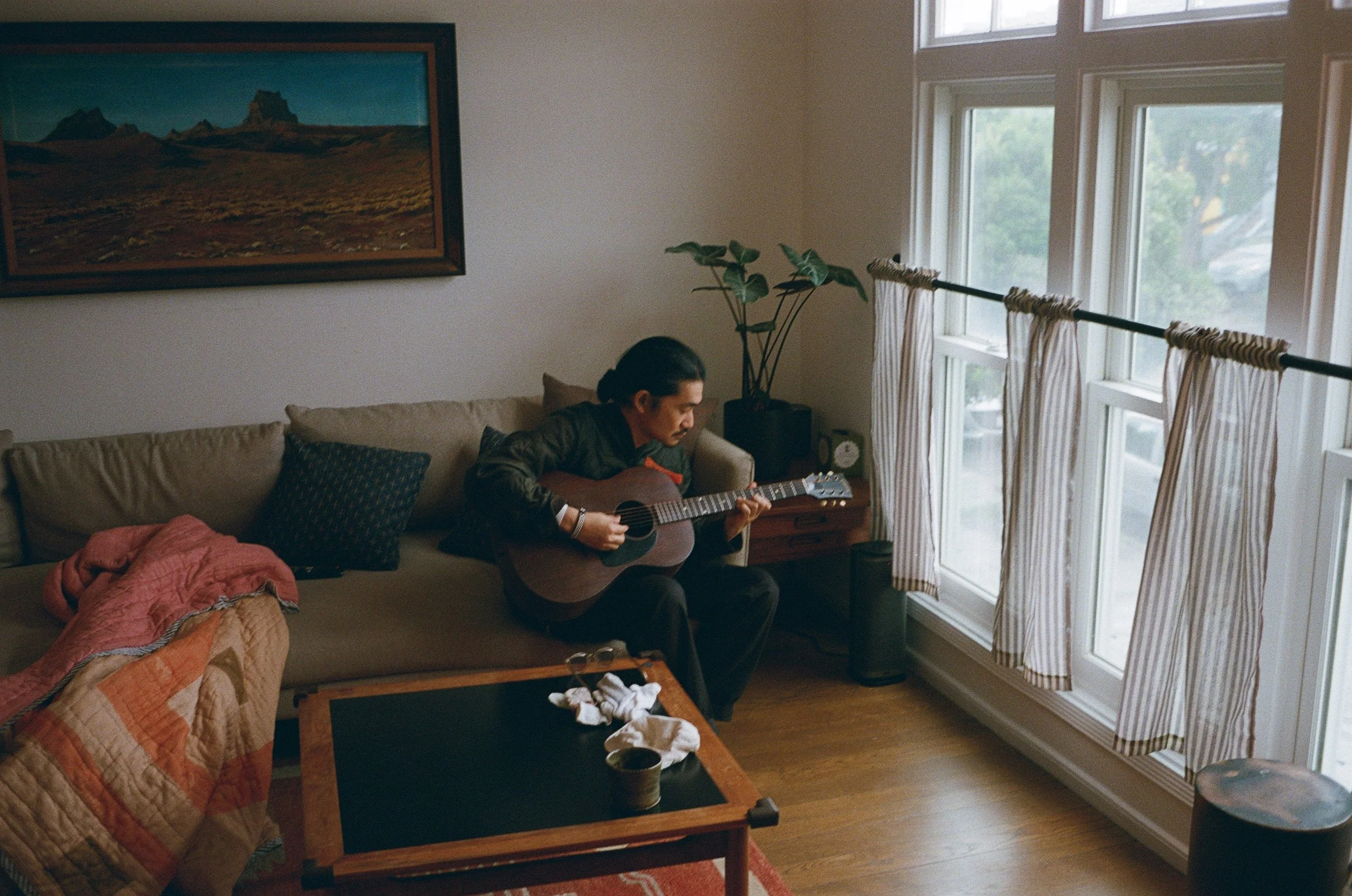 A person with dark hair sitting on a beige couch playing an acoustic guitar in a living room. The room has large windows with striped curtains, a side table with a clock and plant, and a framed landscape painting on the wall.