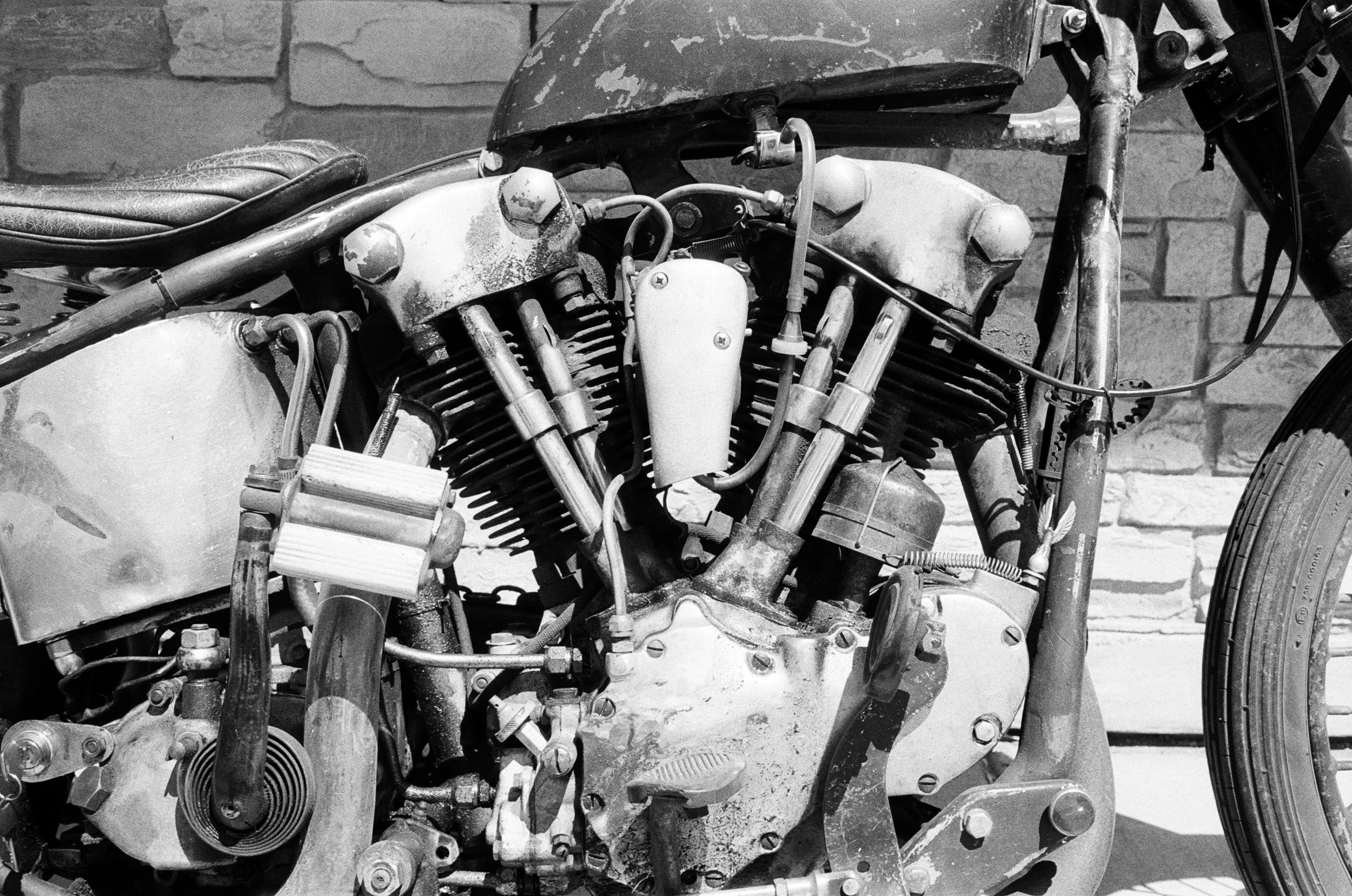 Black and white photograph of an old, mechanical motorcycle engine with various metal parts, pipes, and wires, set against a brick wall background.