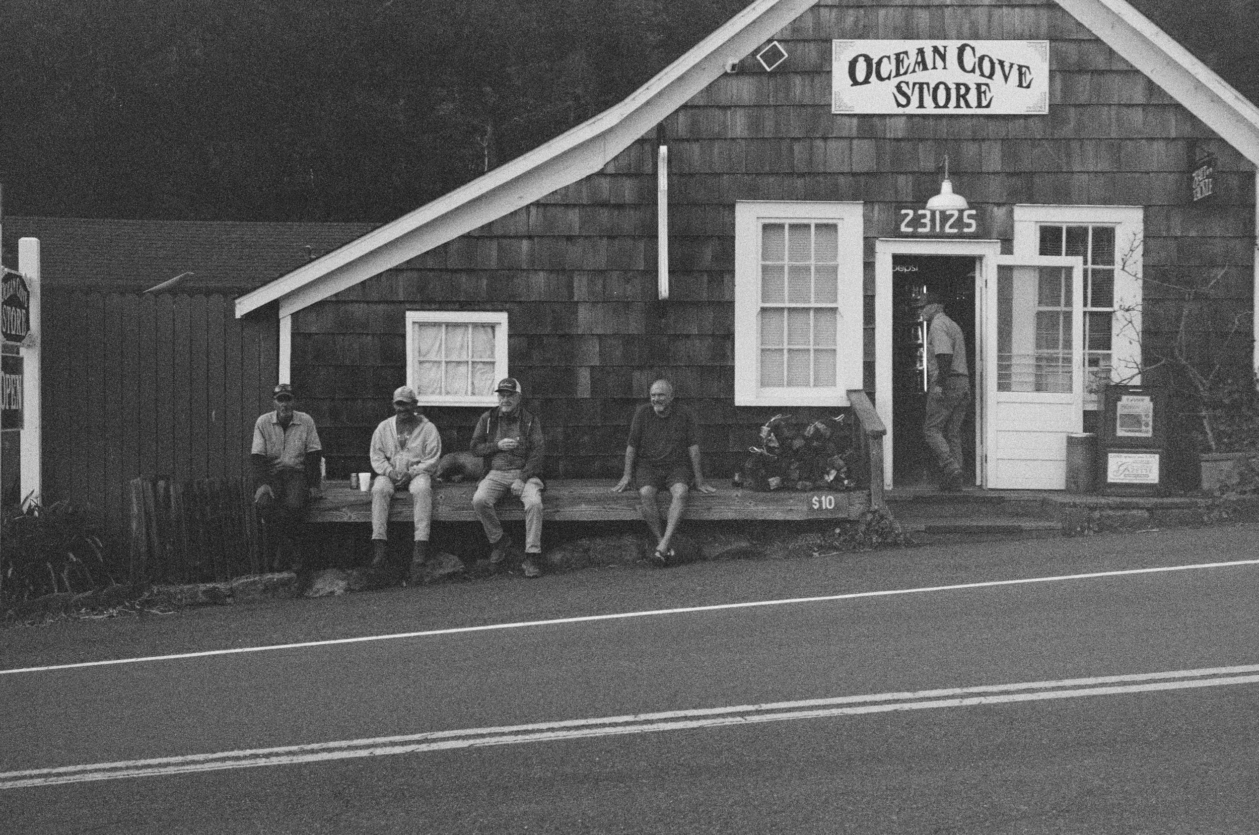 Black-and-white photo of a small wooden store named Ocean Cove Store with four men sitting on a bench outside. The store has an open door, two windows, and a sign displaying the address 2312S. One man is standing inside near the door, and a bicycle is parked against the building. Two signs on each side of the store indicate it is open.
