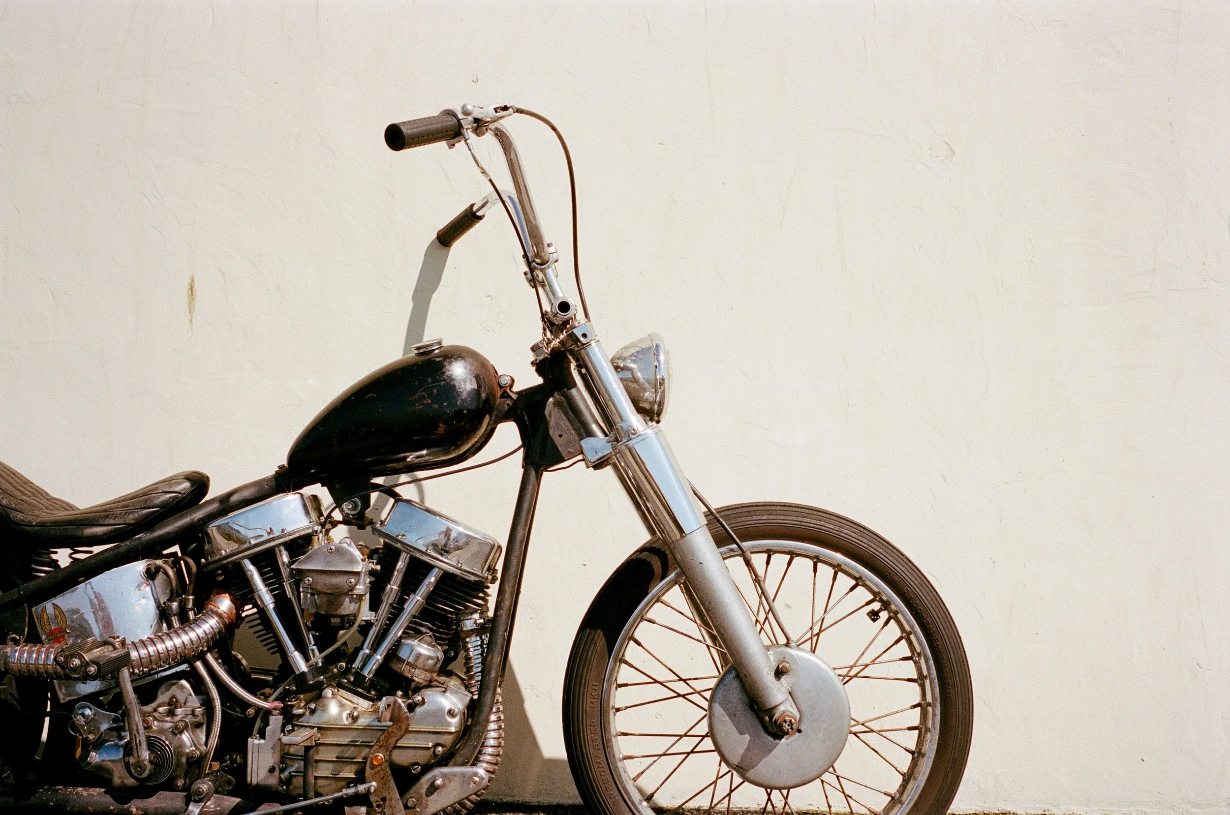 Close-up of a vintage motorcycle with a black fuel tank, chrome engine parts, and a spoked front wheel, positioned against a plain light-colored wall.