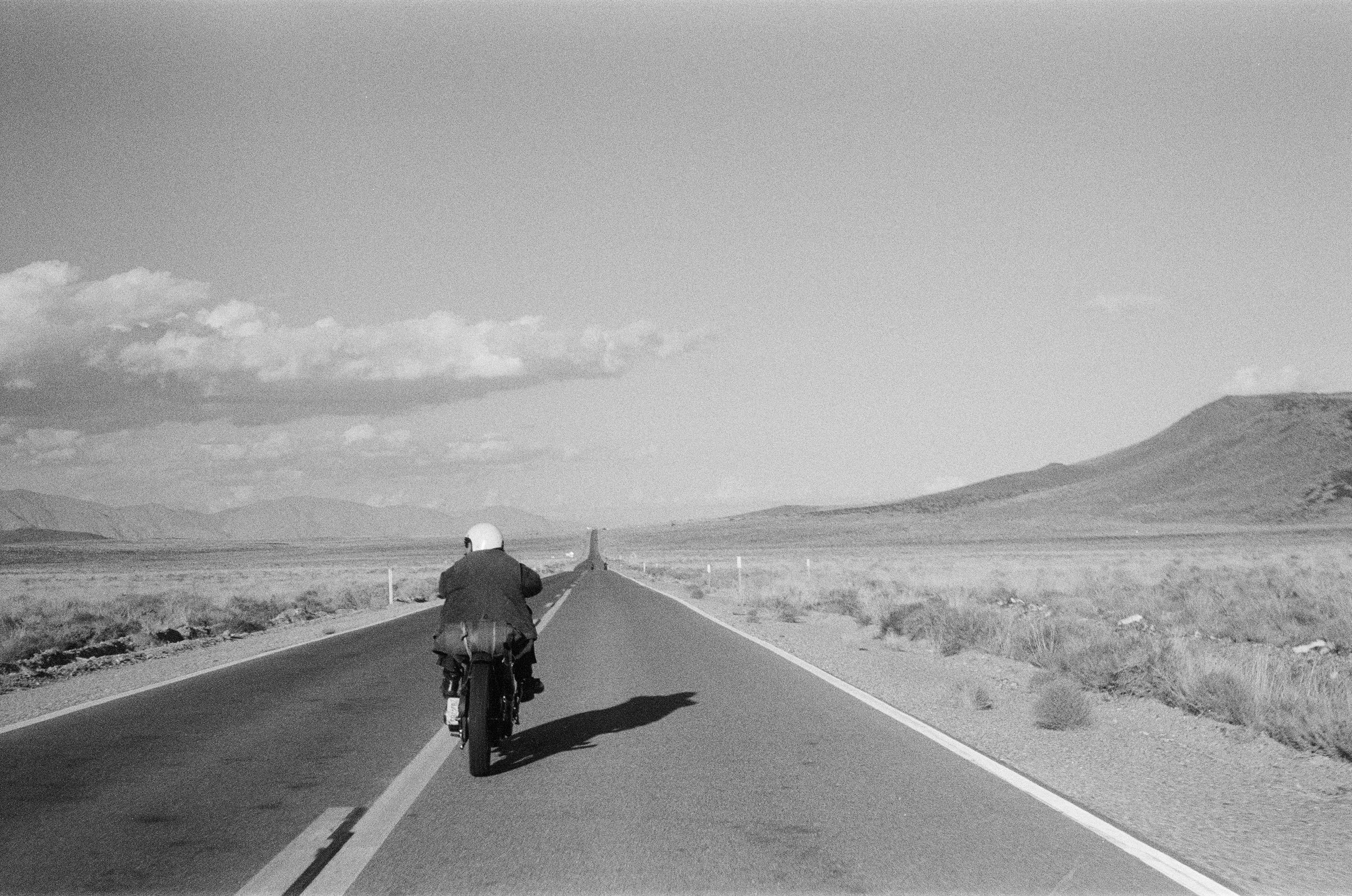 A person wearing a helmet riding a bicycle on an empty road through a flat landscape with mountains in the background and a cloudy sky.
