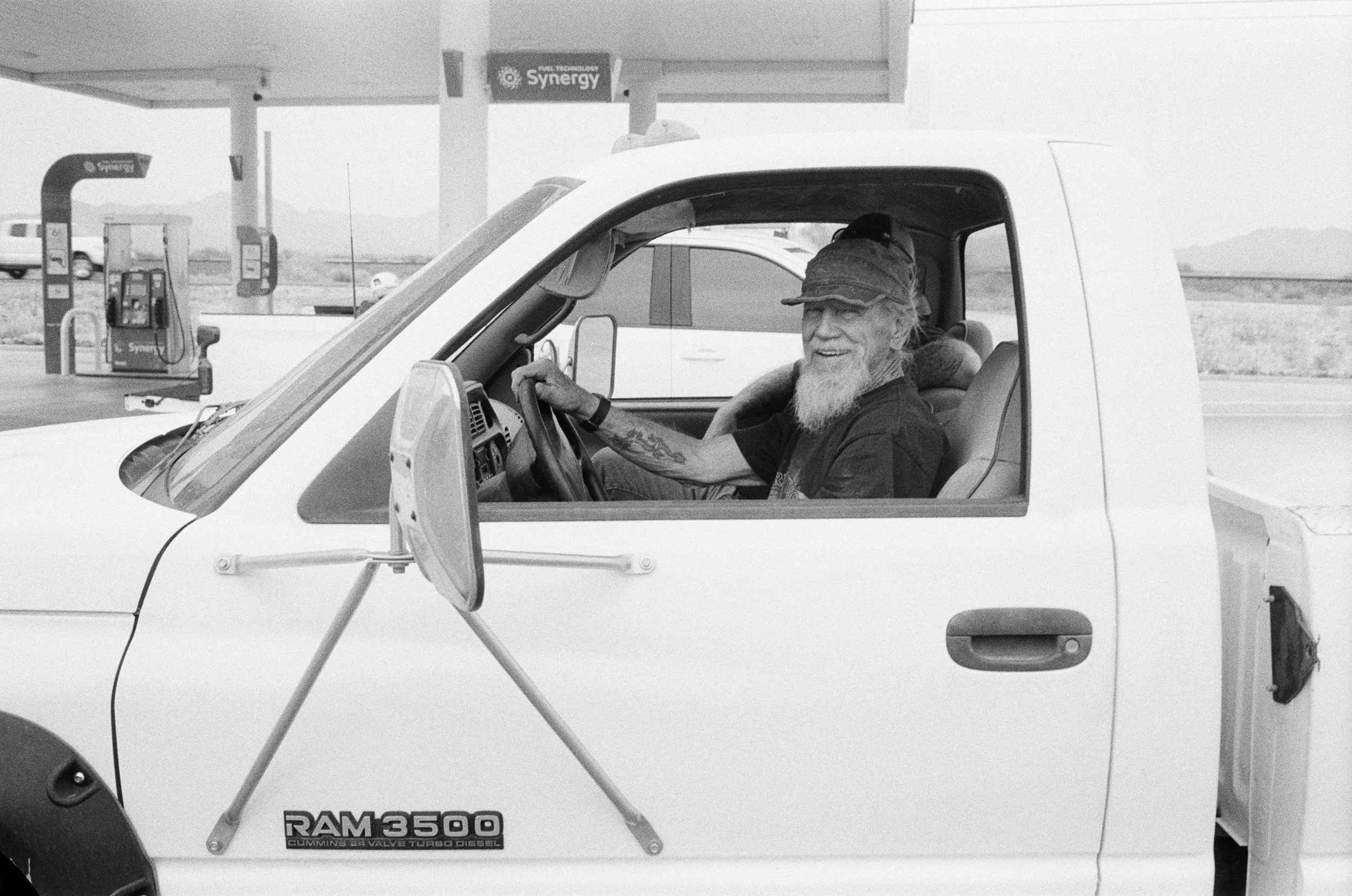 A man with a beard and a hat sitting inside a white Dodge Ram 3500 truck at a gas station, smiling.