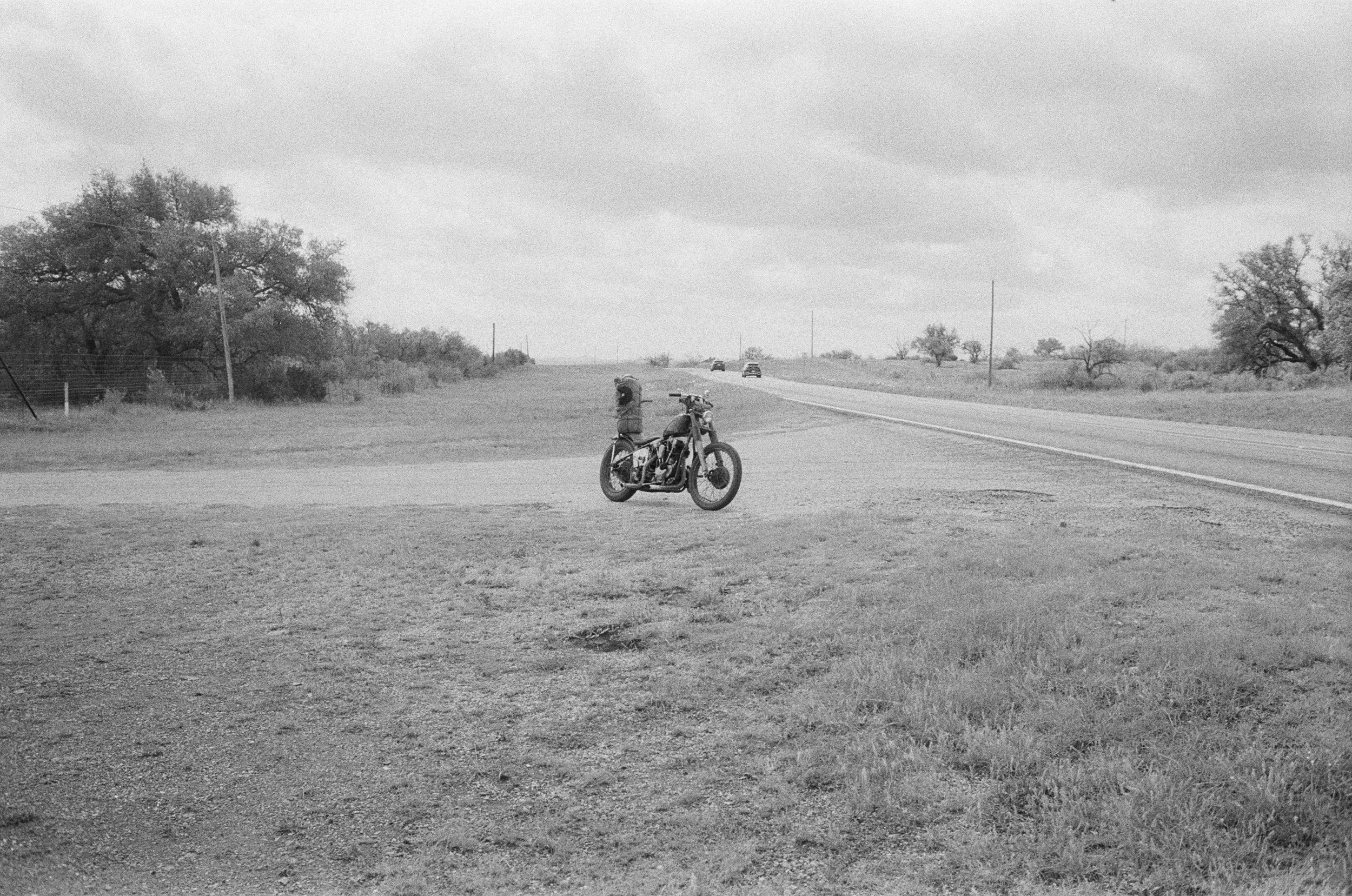 A rural scene with a vintage bicycle parked on the grass near a paved road. In the background, there are trees and a few cars driving on the road under overcast skies.