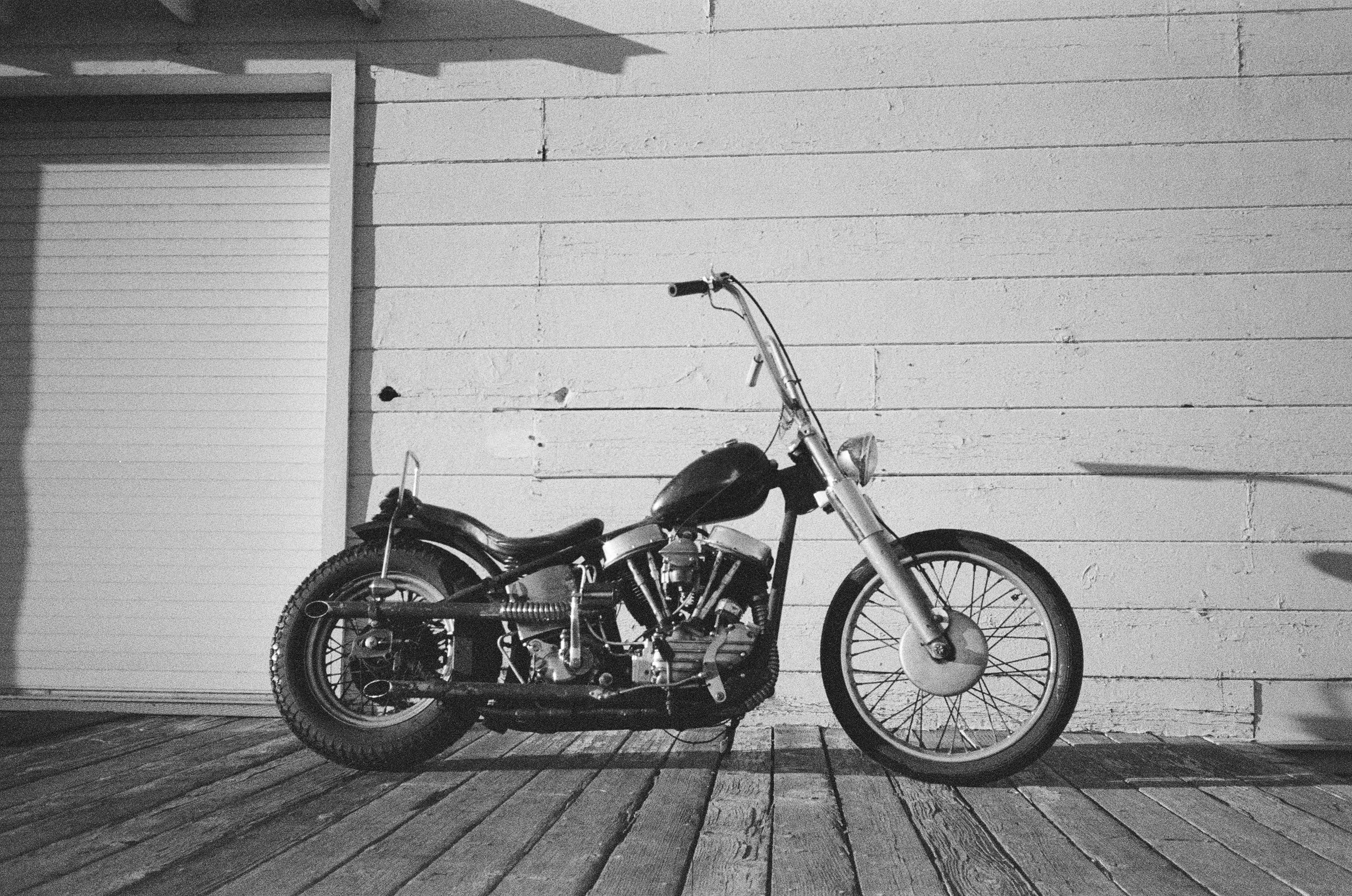 A black motorcycle parked on a wooden porch in front of a white painted wooden wall.