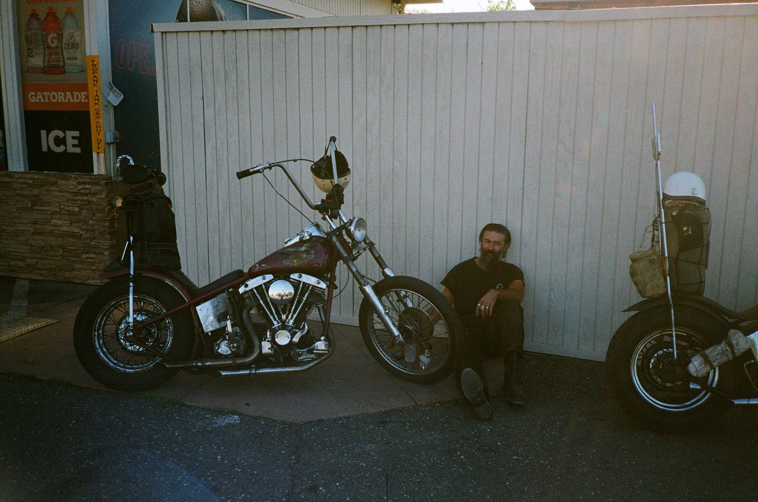 A man with a beard sitting on the ground next to a parked motorcycle with a white wall background. There is a large bag on a pole to the right and a person wearing a helmet and backpack standing nearby. A Gatorade ice cooler is partially visible on the left side.