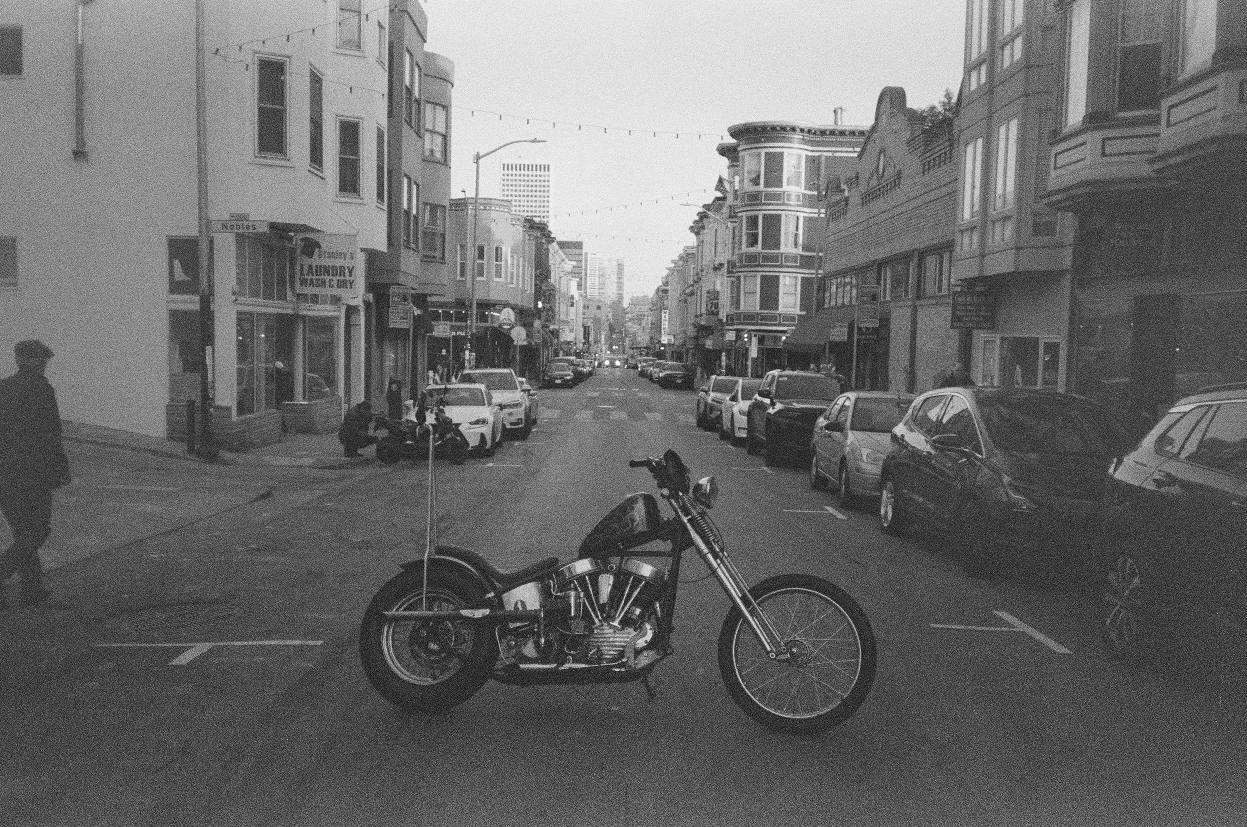 Black and white street scene with parked cars, buildings on both sides, and a motorcycle in foreground. A person is walking on the sidewalk on the left, with signage indicating a laundry service. Overhead string lights hang across the street.