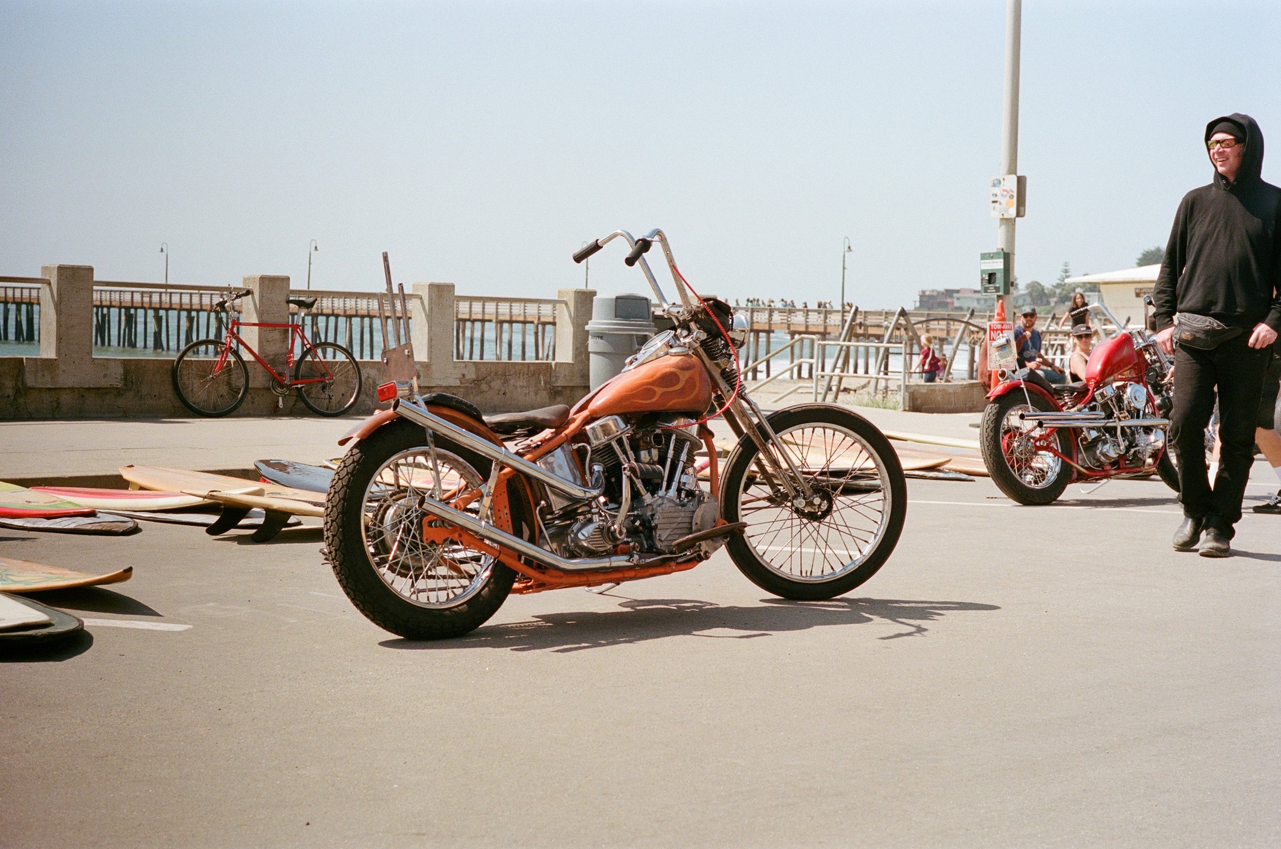 A vintage motorcycle with flame detailing parked on a concrete surface, with surfboards on the ground nearby. In the background, people are sitting and standing near the pier and ocean on a foggy day. A man in a black hoodie and sunglasses is smiling and walking near the motorcycles.