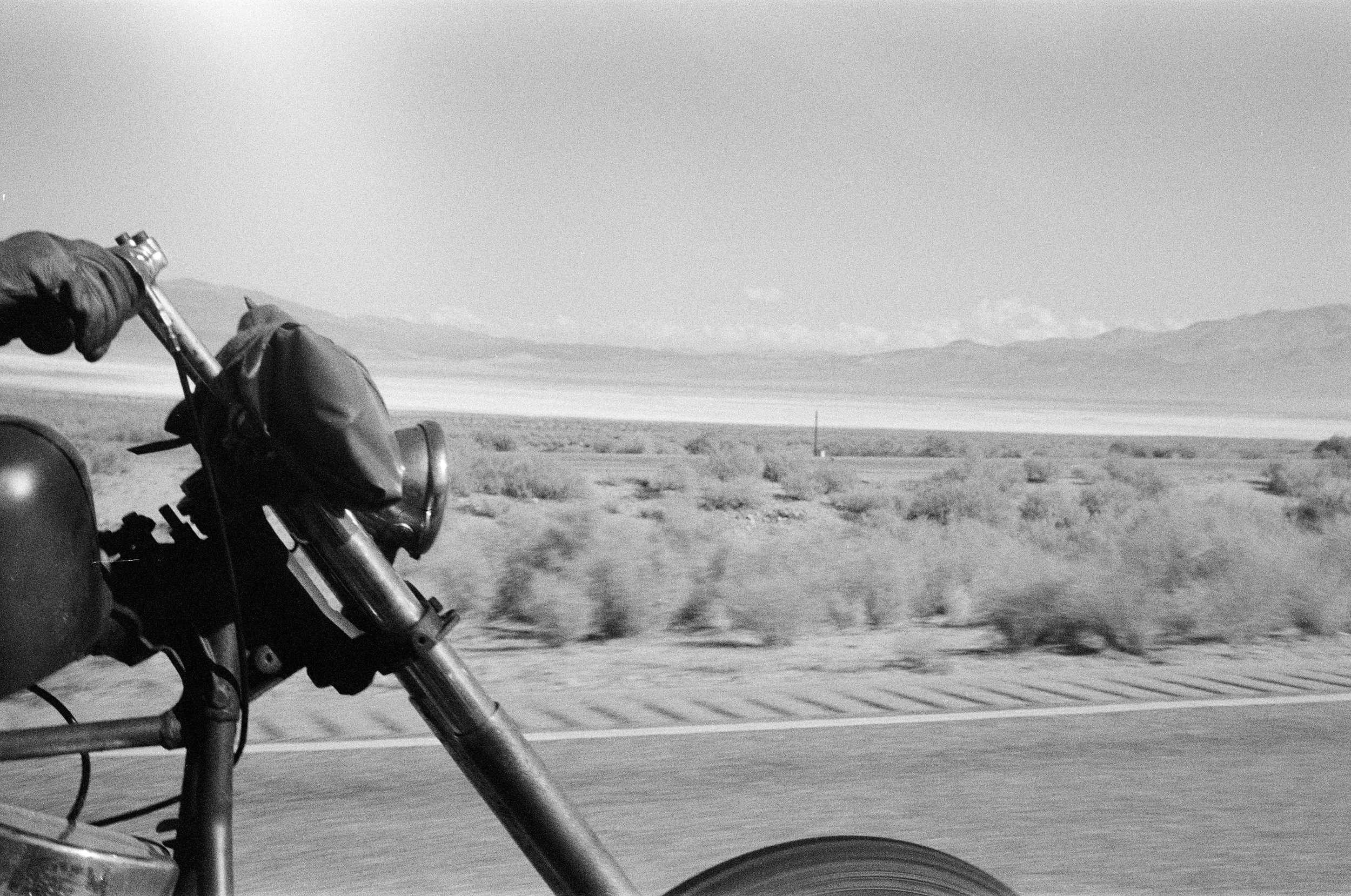 Close-up view of a motorcycle front wheel and handlebars in motion on an open road with a desert landscape and mountains in the distance.