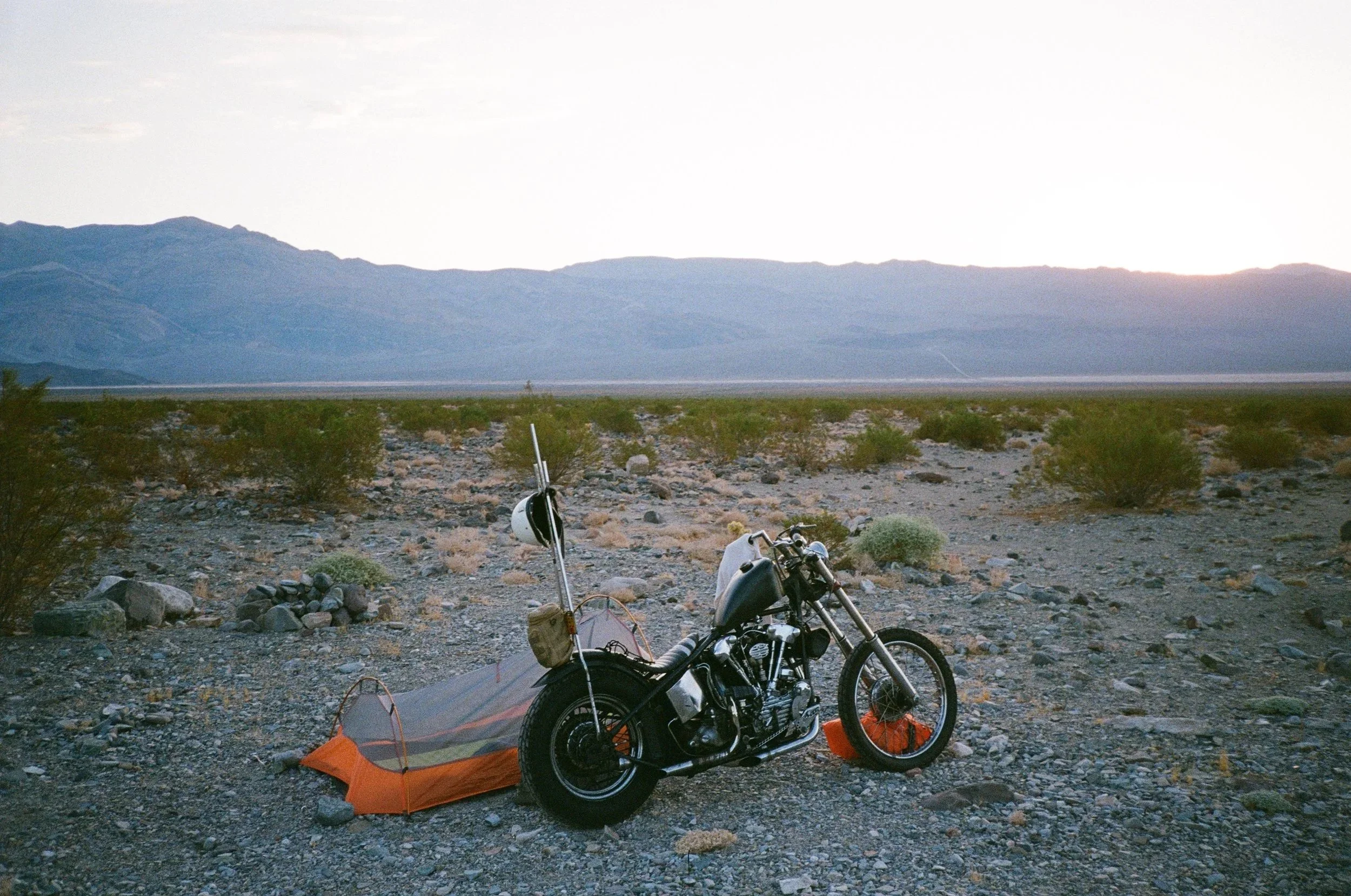 A motorcycle parked next to a tent in a desert landscape with mountains in the background during sunset.