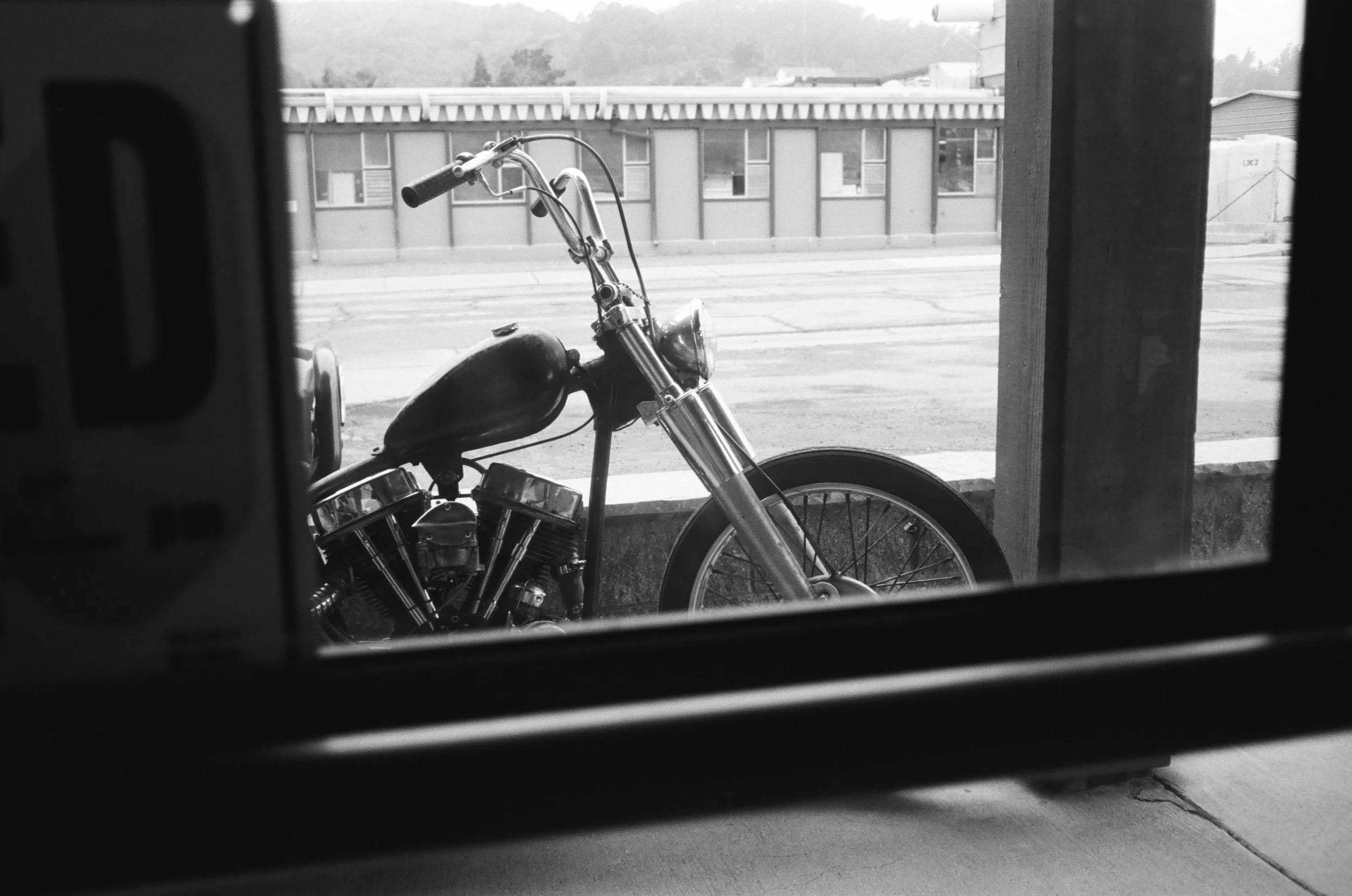 A black and white photo of a motorcycle seen through an open garage door or window, with the street visible in the background.