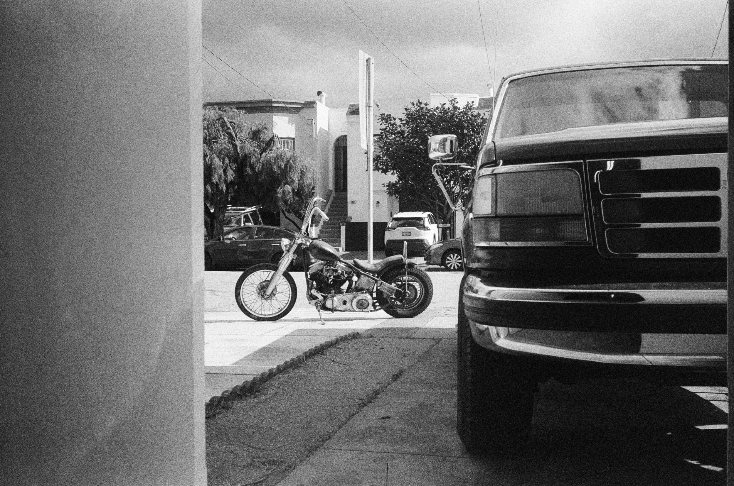 Black and white photo of a street scene. A motorcycle is parked on the sidewalk, and several cars, including a large vehicle, are visible. There are trees and residential buildings in the background. The photo is partially framed by a wall on the left side.