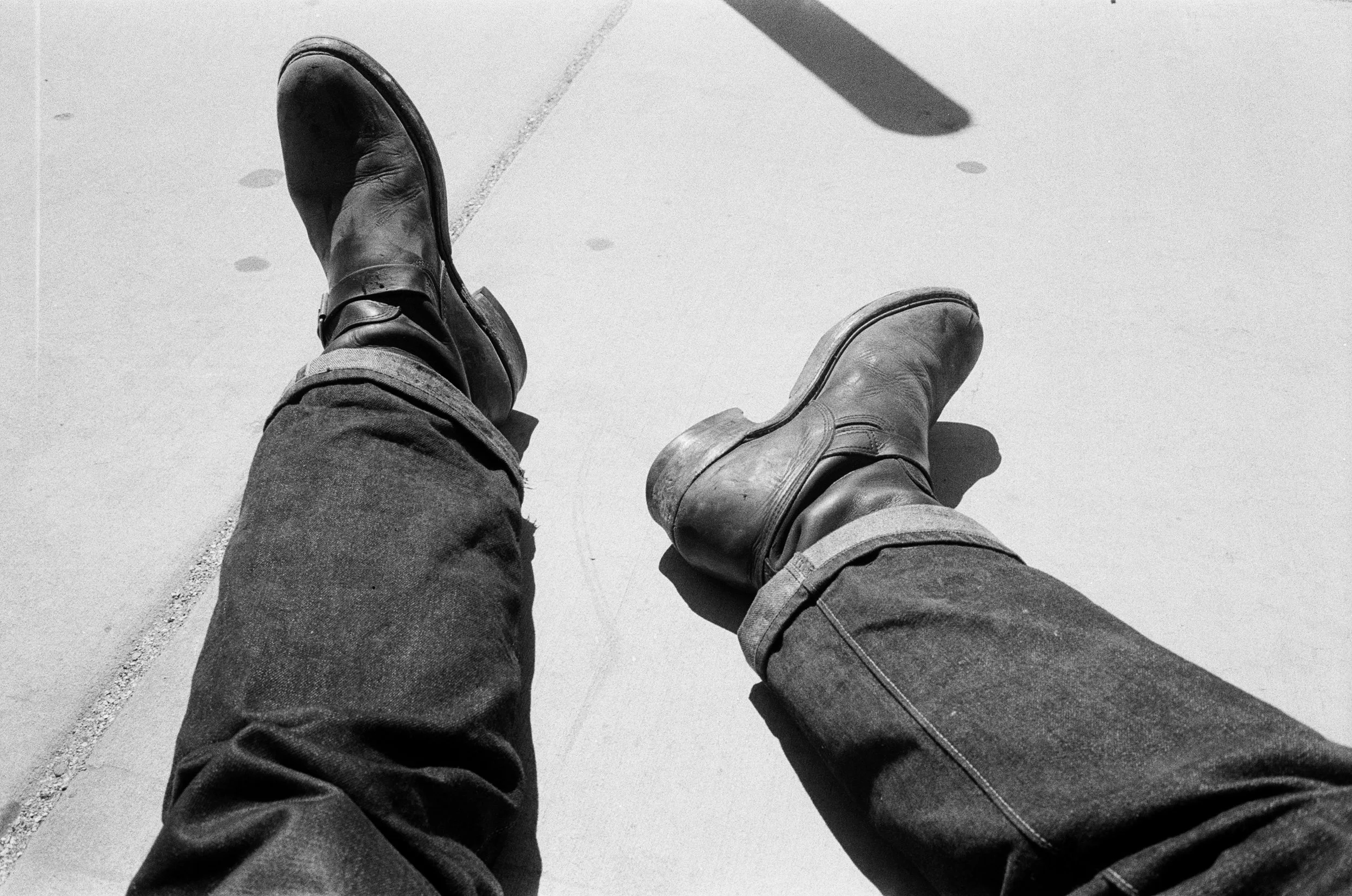 A black and white photograph of a person's legs and feet, wearing worn leather boots and jeans, sitting on a concrete surface with their legs stretched out.