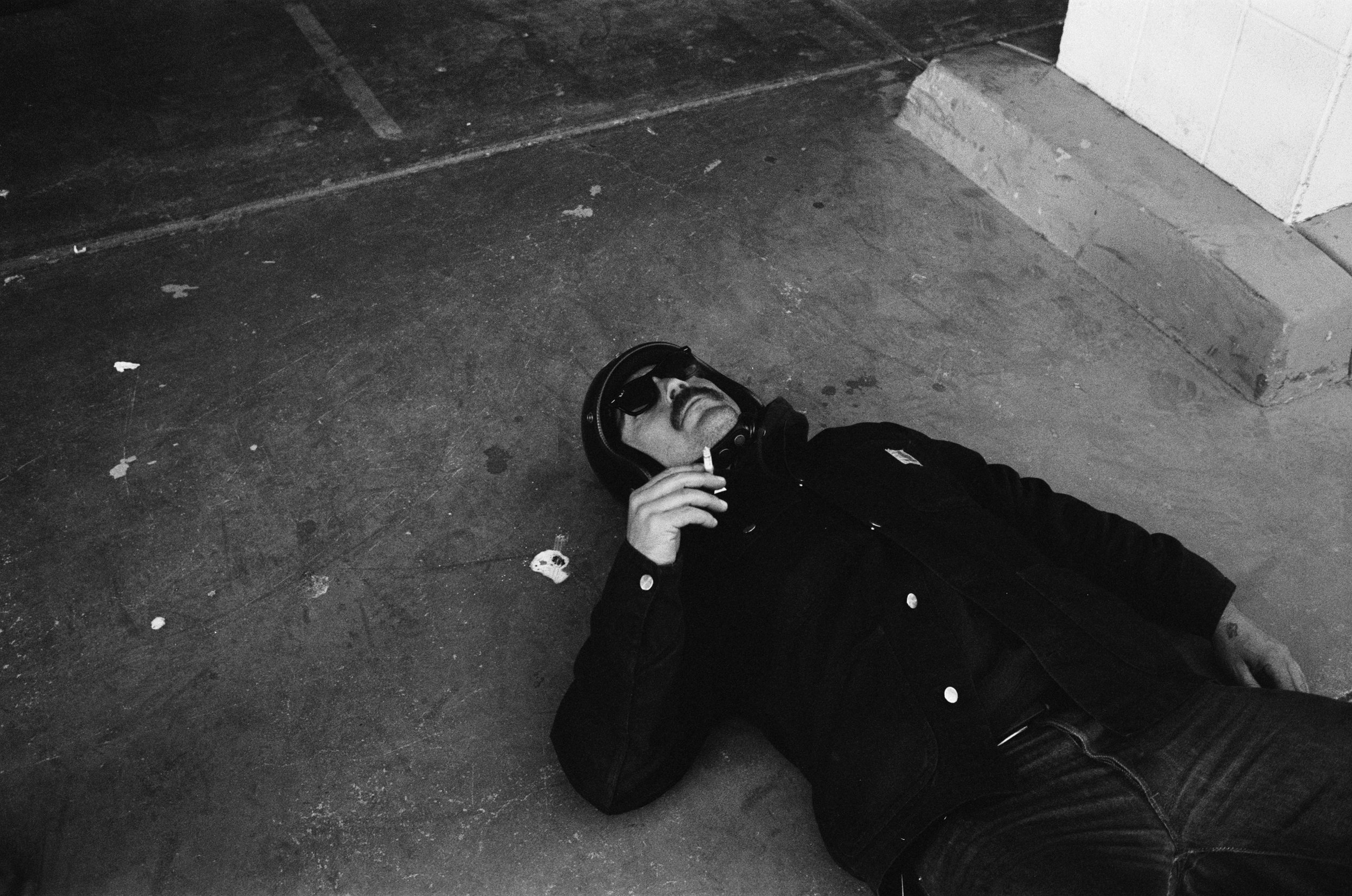 A person lying on the floor wearing black clothing and headphones, holding a cigarette near their face, in a dimly lit setting with a concrete or tiled floor and white tiled wall.