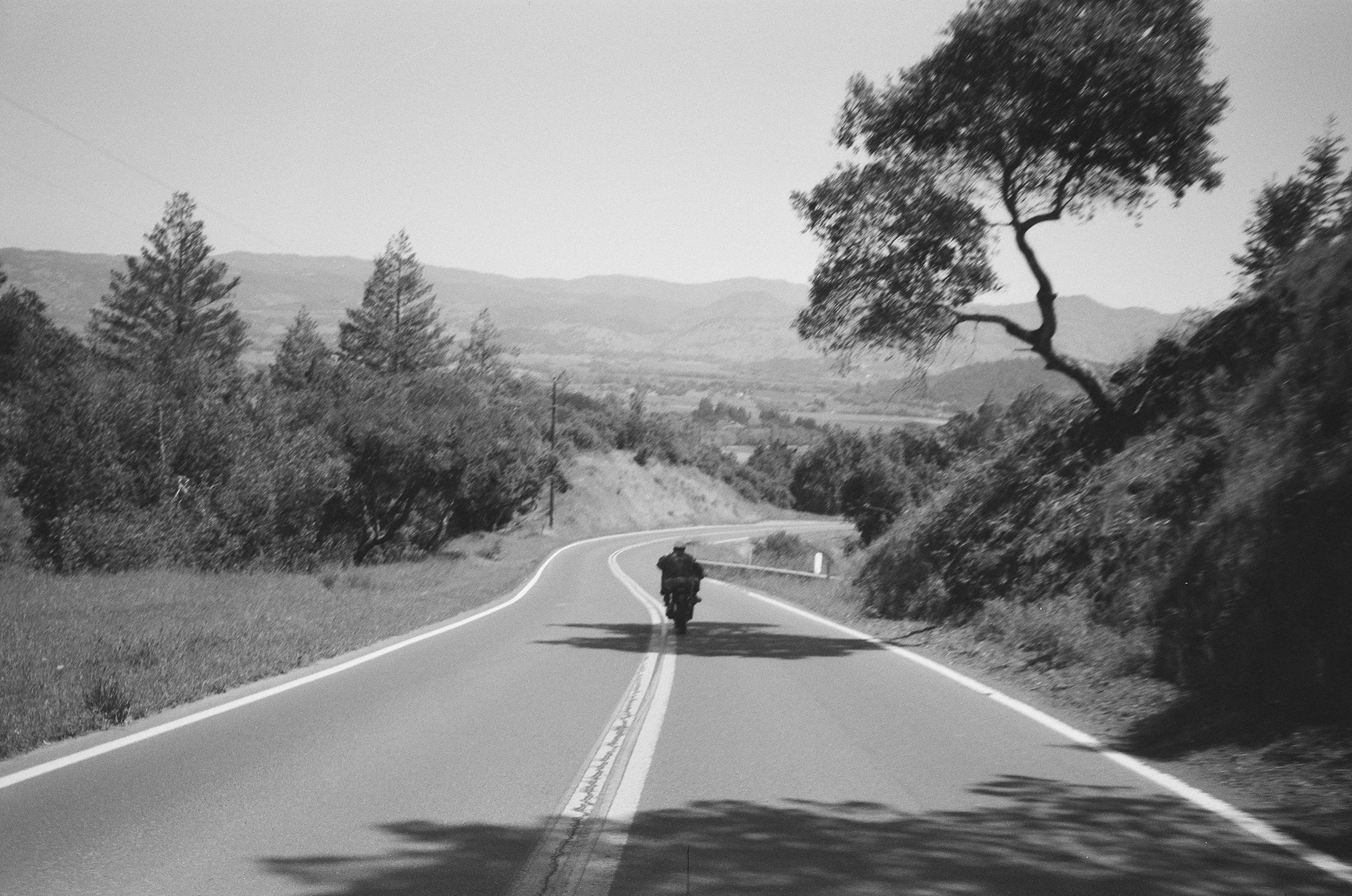 A black and white photo of a person riding a bicycle on a winding road with trees and hills in the background.