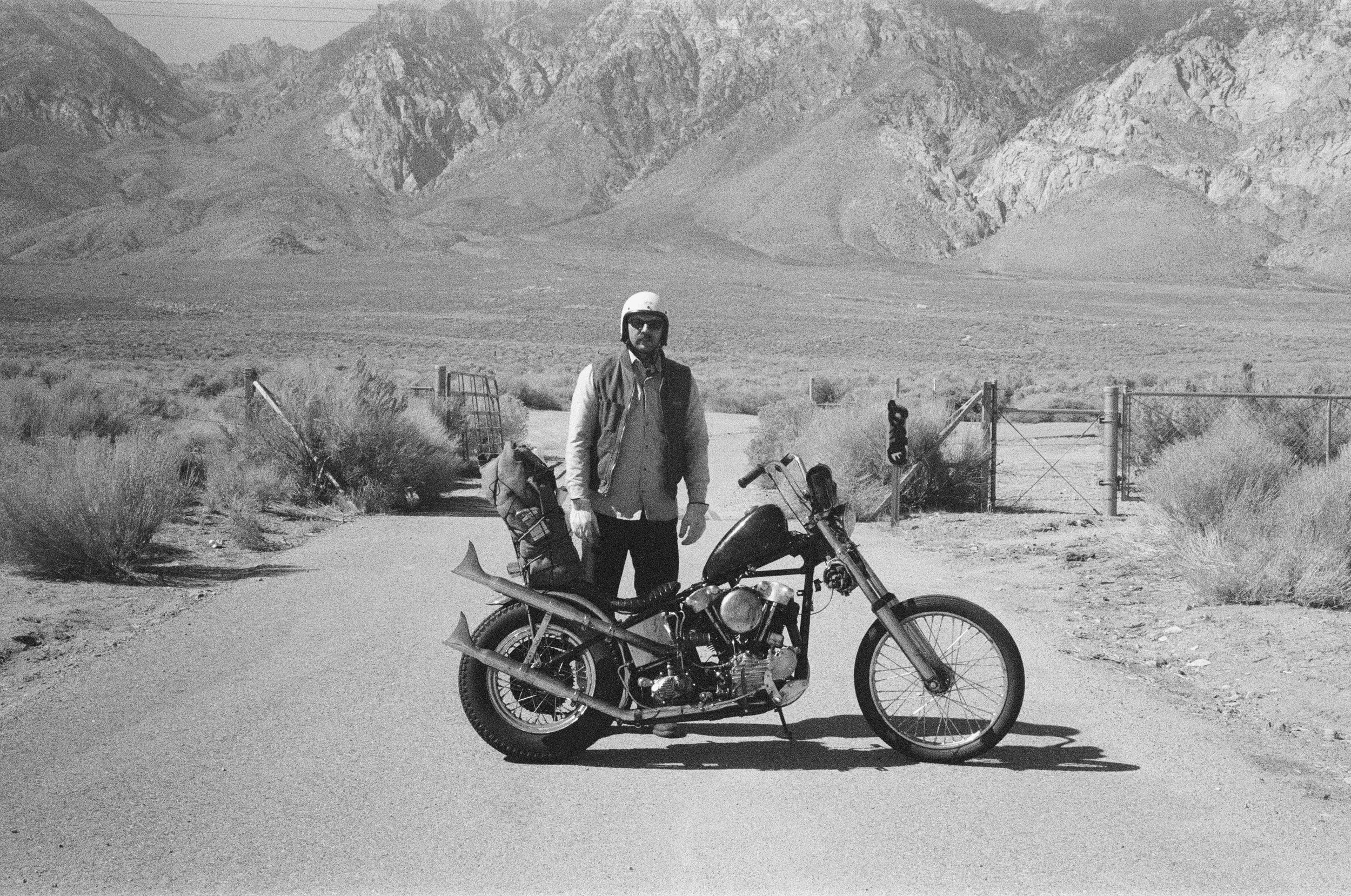 A man wearing a helmet and sunglasses standing beside a black motorcycle in a desert landscape with mountains in the background.