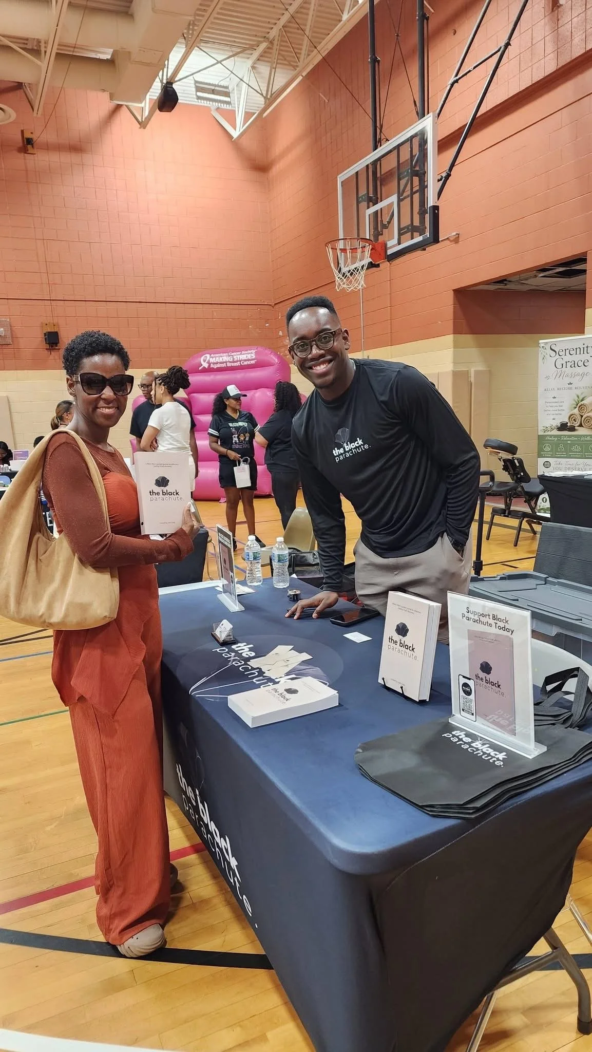 A woman with sunglasses and a brown jacket holding a brochure stands next to a man at a booth with books and items promoting 'the black parachute' at an indoor event in a gymnasium. The man is smiling and leaning on the table. Background shows other 