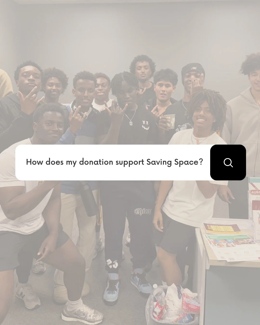 Group of young men posing together at an indoor event, some making hand signs, with a table holding flyers and a donation box nearby.