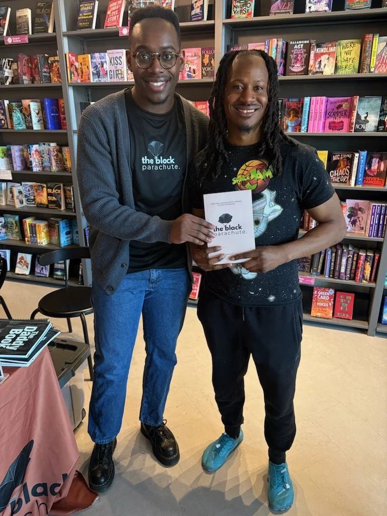 Two men smiling and holding a book inside a bookstore with shelves of colorful books in the background.