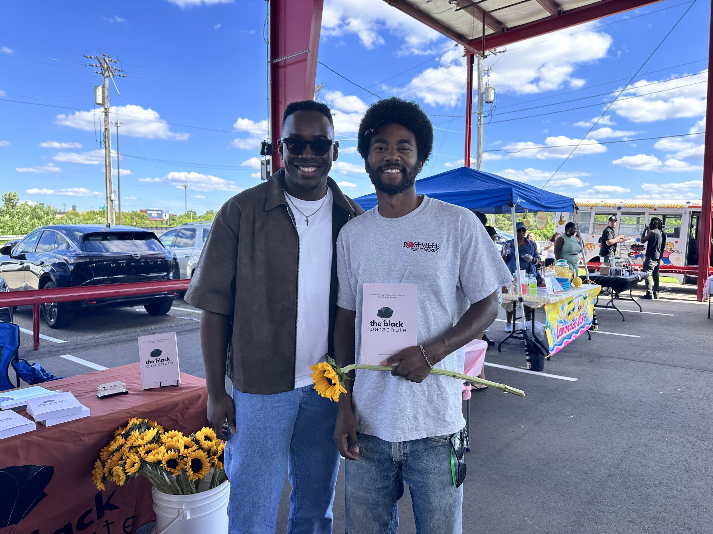 Two men standing together outdoors during a daytime event, holding a sunflower, with tables and tents in the background, and a clear sky with some clouds.