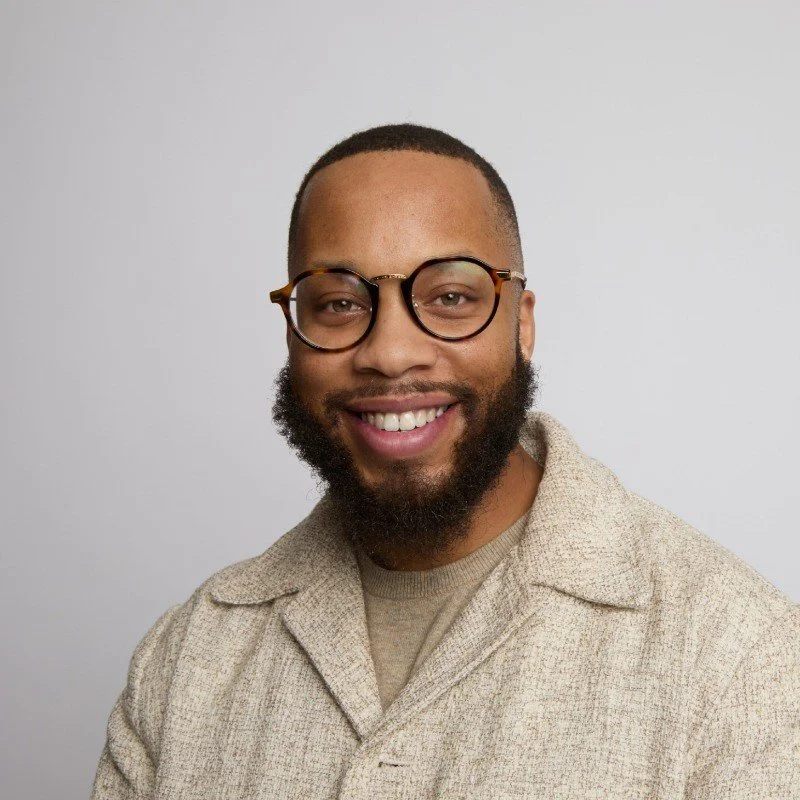 A smiling man with glasses, a beard, and short hair, wearing a beige collared shirt, standing against a plain gray background.