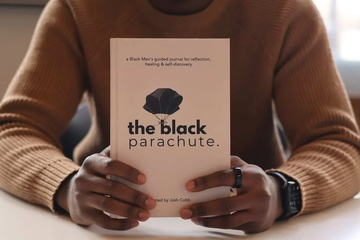 Person wearing a tan sweater holding a book titled "the black parachute" with a black parachute illustration on the cover.