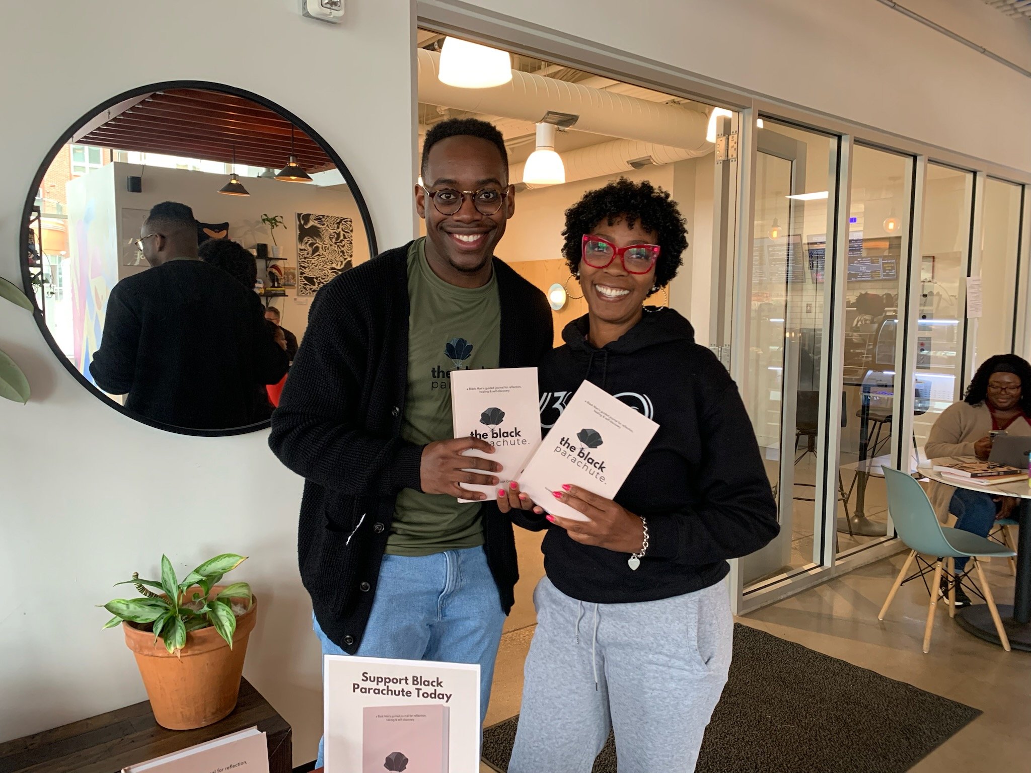 Two people smiling and holding flyers at a support Black parachute event, standing indoors near a mirror and a plant.