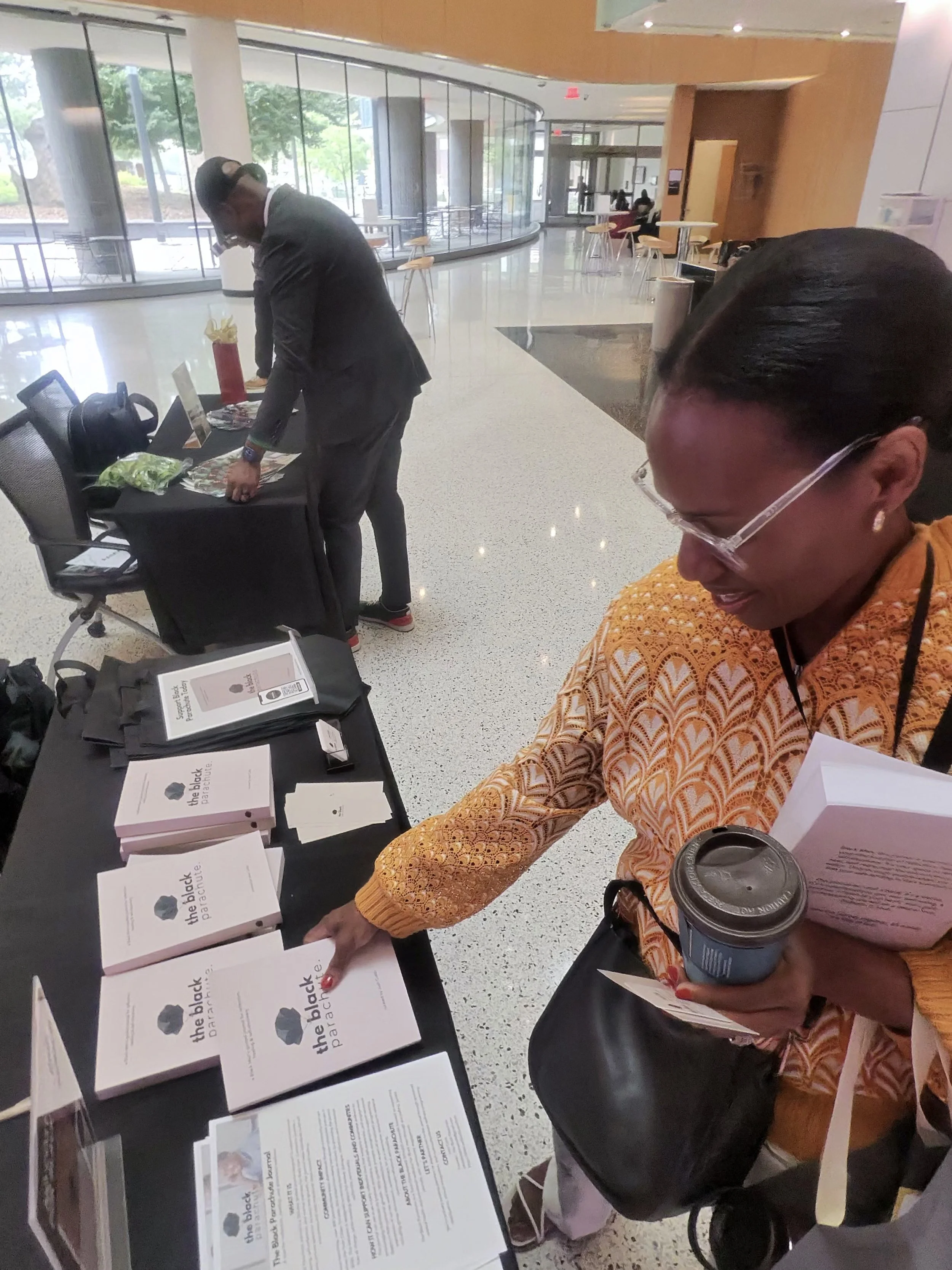 A woman in an orange patterned top is reaching for printed materials on a table, holding a coffee cup, with multiple copies of a booklet titled 'the black parachute' displayed on the table. A man in a dark suit is standing nearby, looking at the materials, in a lobby or conference area with large glass windows and tables.