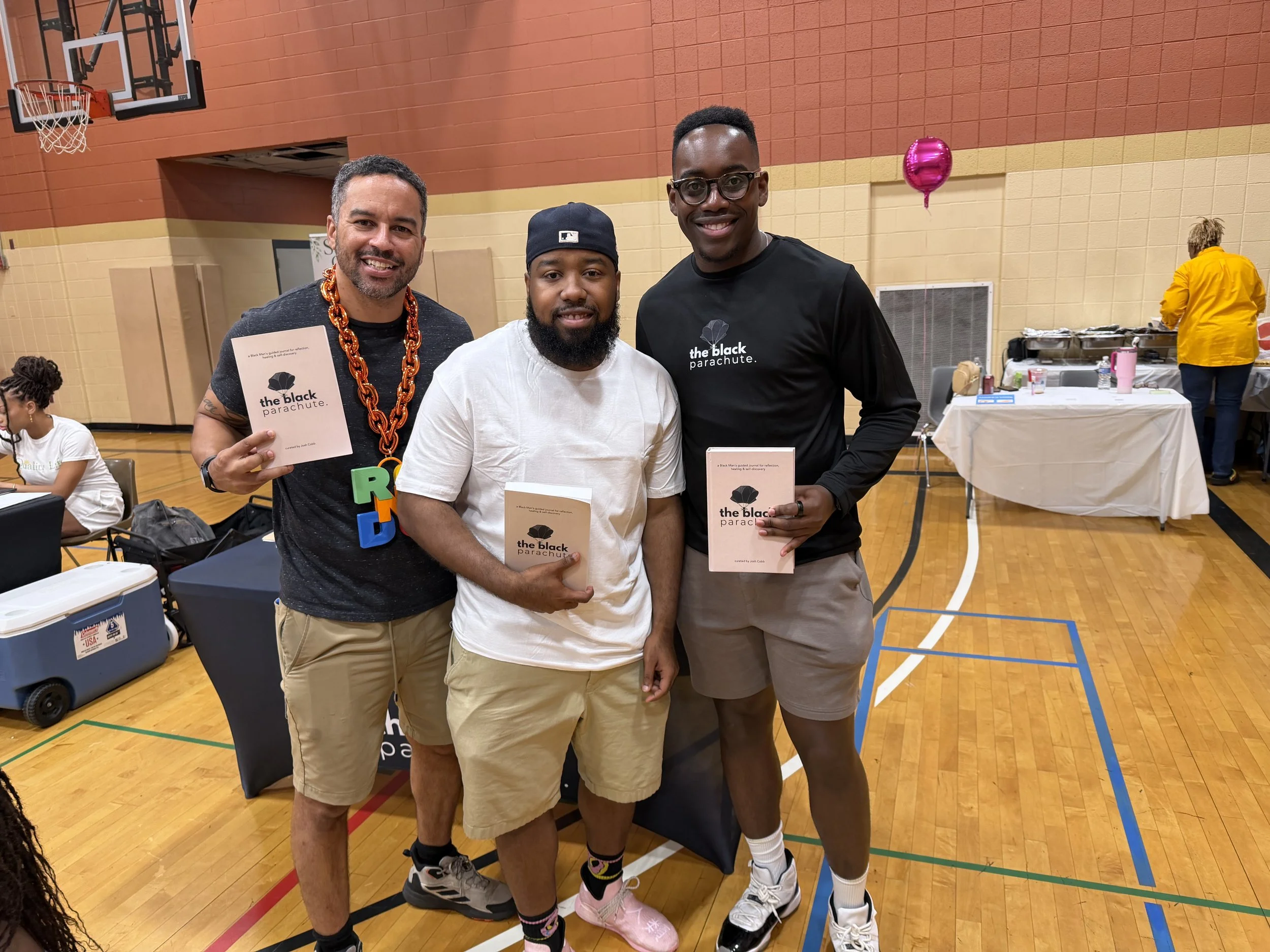 Three men standing inside a gymnasium, holding books titled "the black parachute." They are smiling and wearing casual clothing, with the middle man wearing a black cap and beard. In the background, there are tables, people, a pink balloon, and a wom