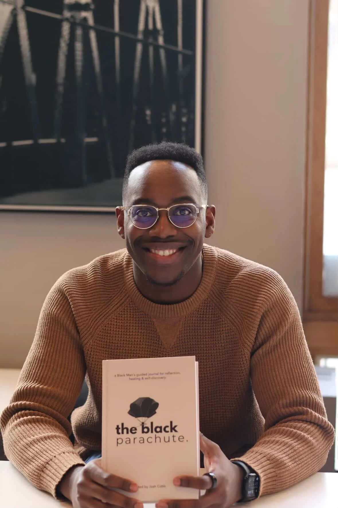 A smiling man with glasses and a brown sweater sitting at a table, holding a book titled 'the black parachute' by Joël Cacou.