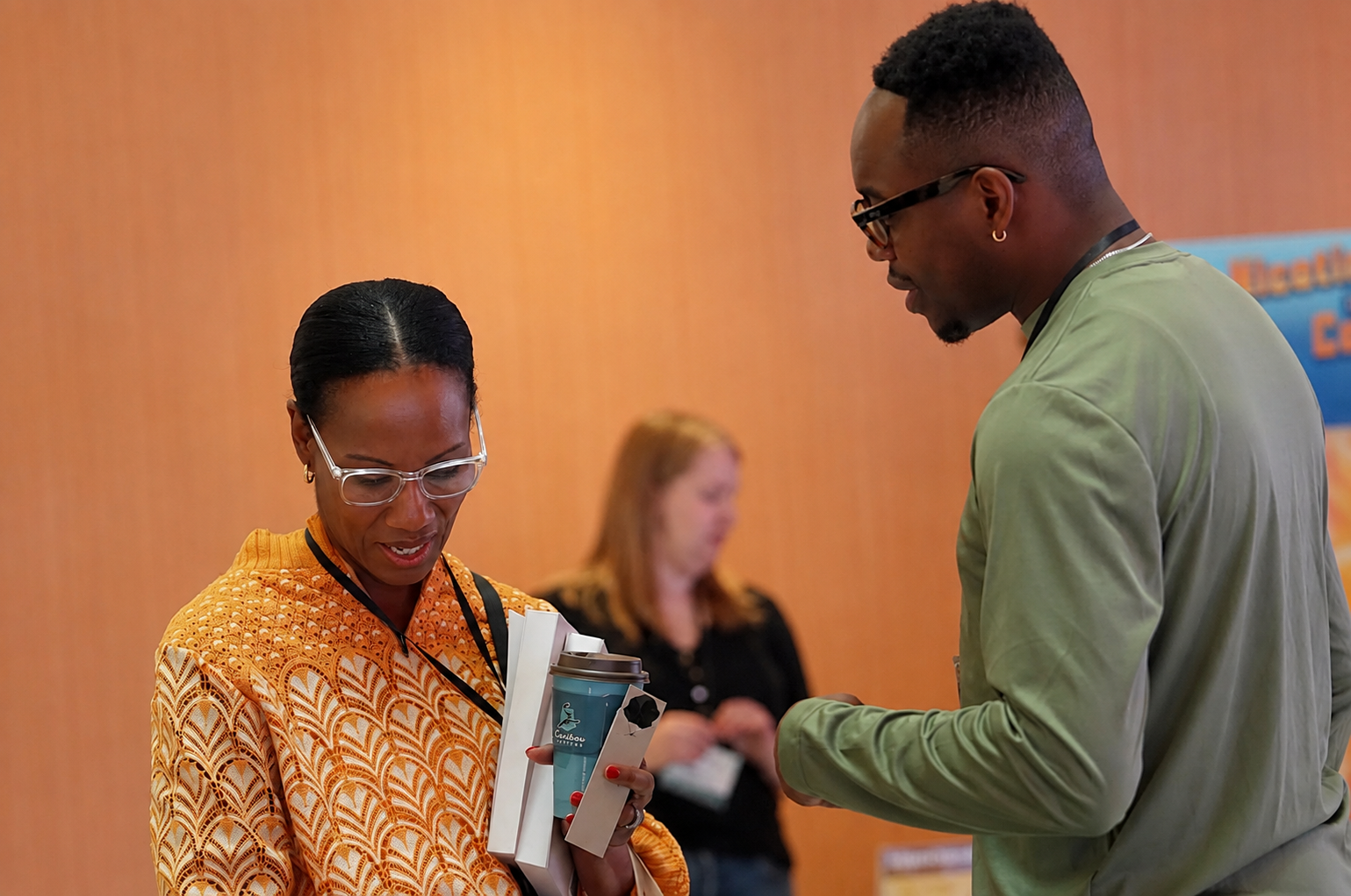 A woman with glasses in an orange patterned top looks at her items including a notebook, a coffee cup, and possibly a brochure, while a man in a green jacket engages in conversation with her at an indoor event.