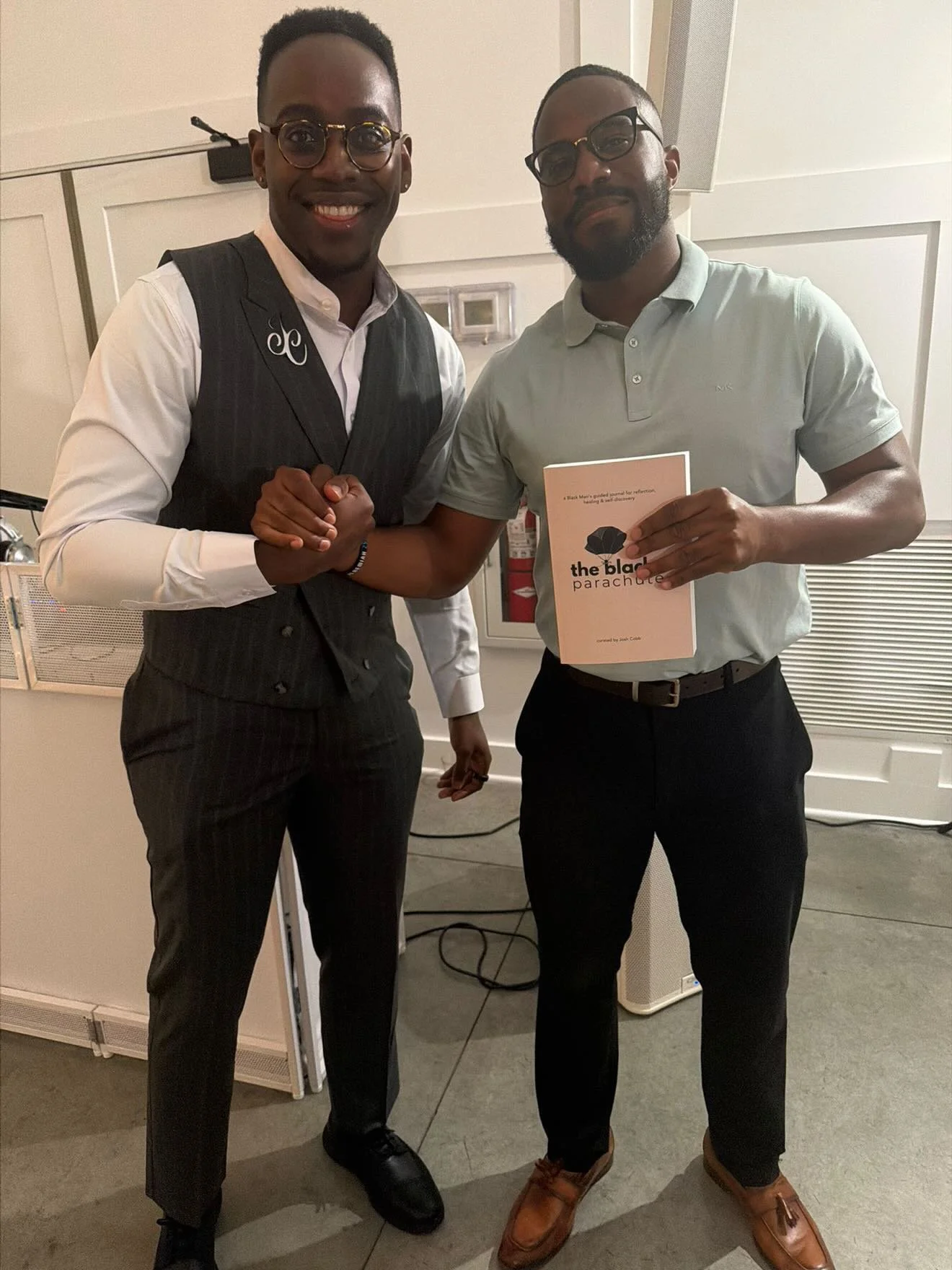 Two men, one in a pinstripe vest and pants and the other in a light green polo and dark slacks, stand together shaking hands and smiling for the camera. The man on the right holds a booklet titled 'the black parachute' in his left hand. Both are wear