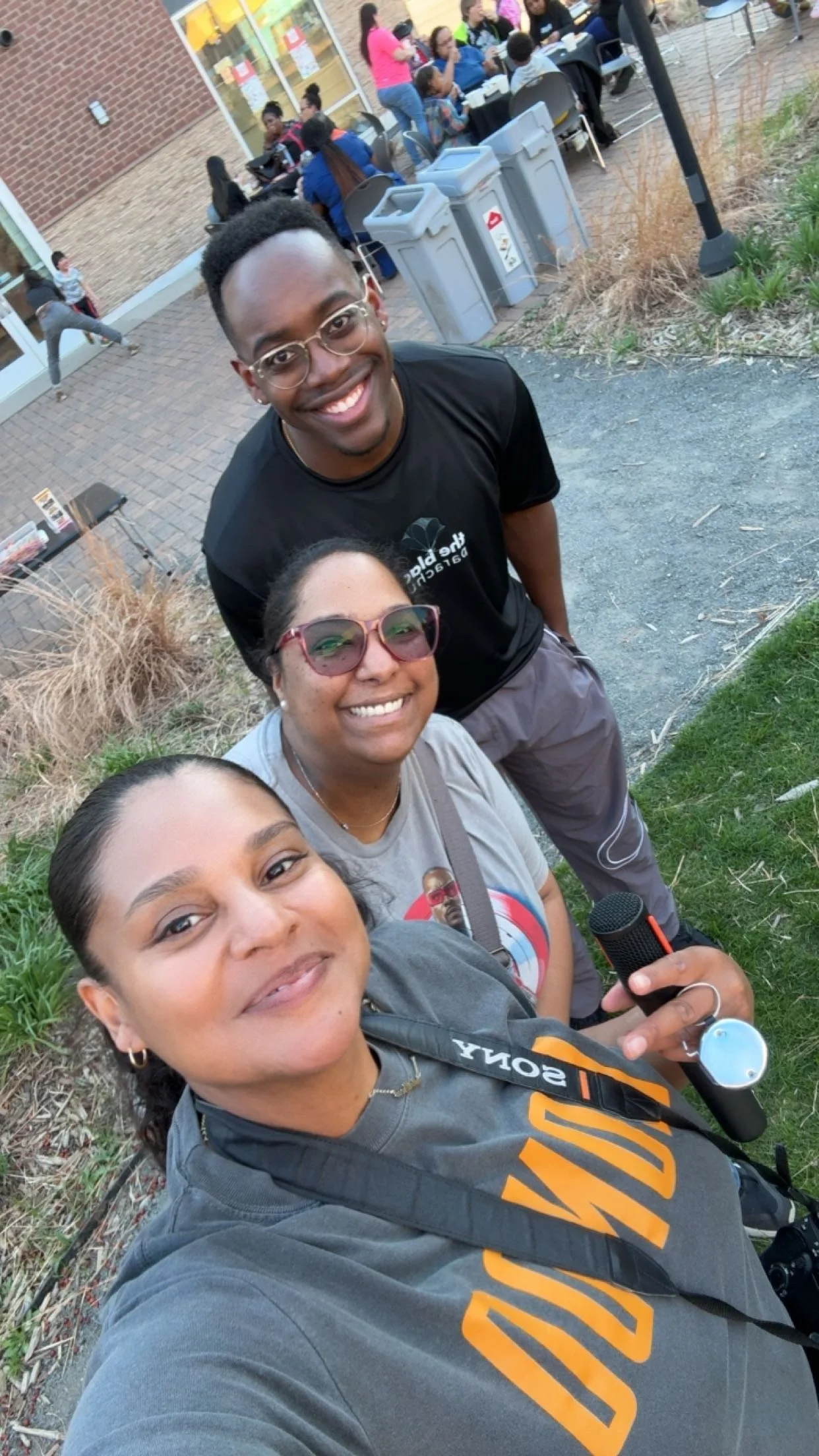 A group of four friends taking a selfie outdoors. Three women and one man smiling at the camera, with a grassy area and a build of people sitting at tables in the background.