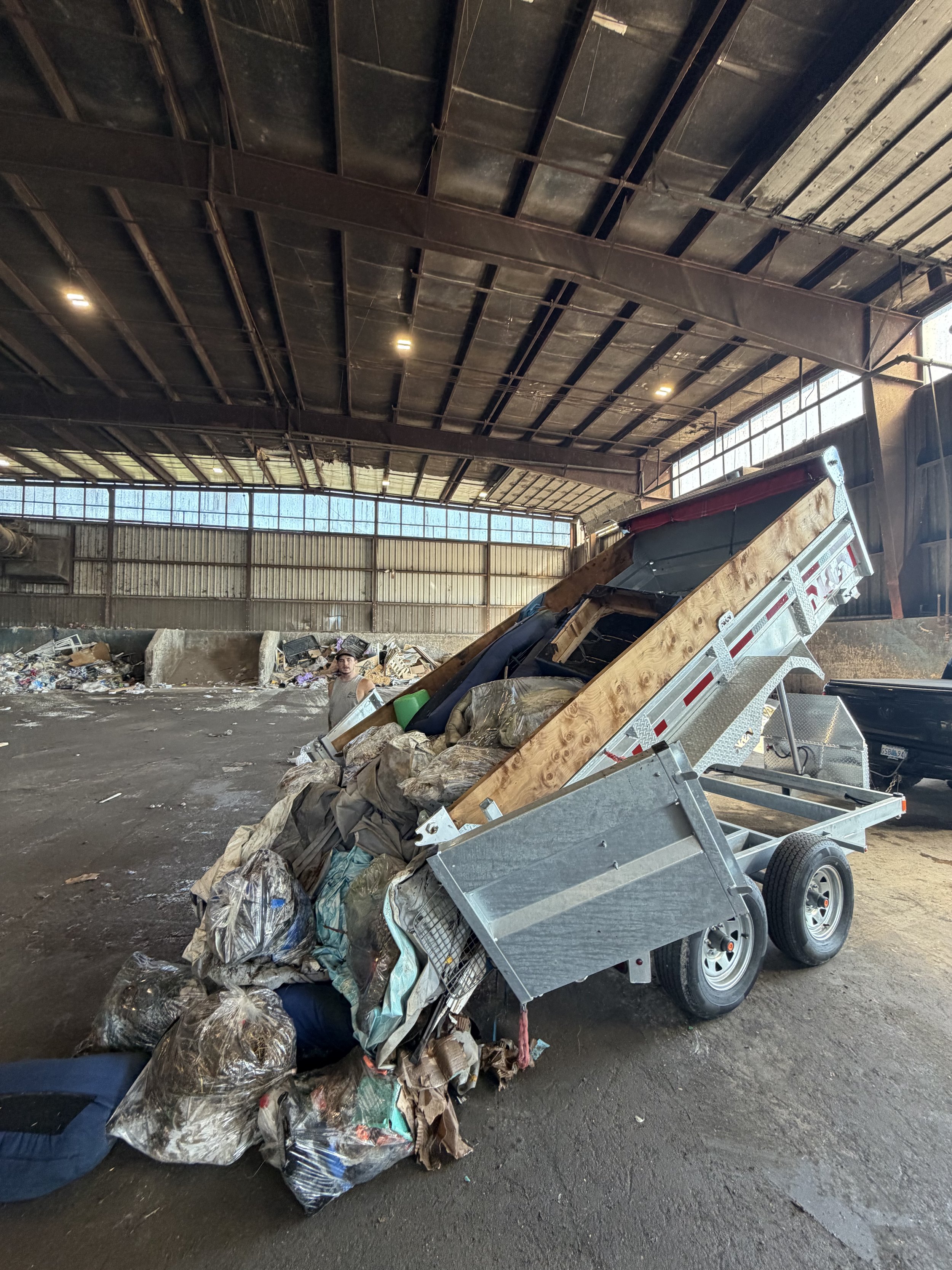 An industrial warehouse with a high ceiling, metal beams, and windows along the upper part of the walls. A dump trailer filled with trash, including bags and debris, is tipped over on the gravel floor. A person is visible near the trash, looking towa