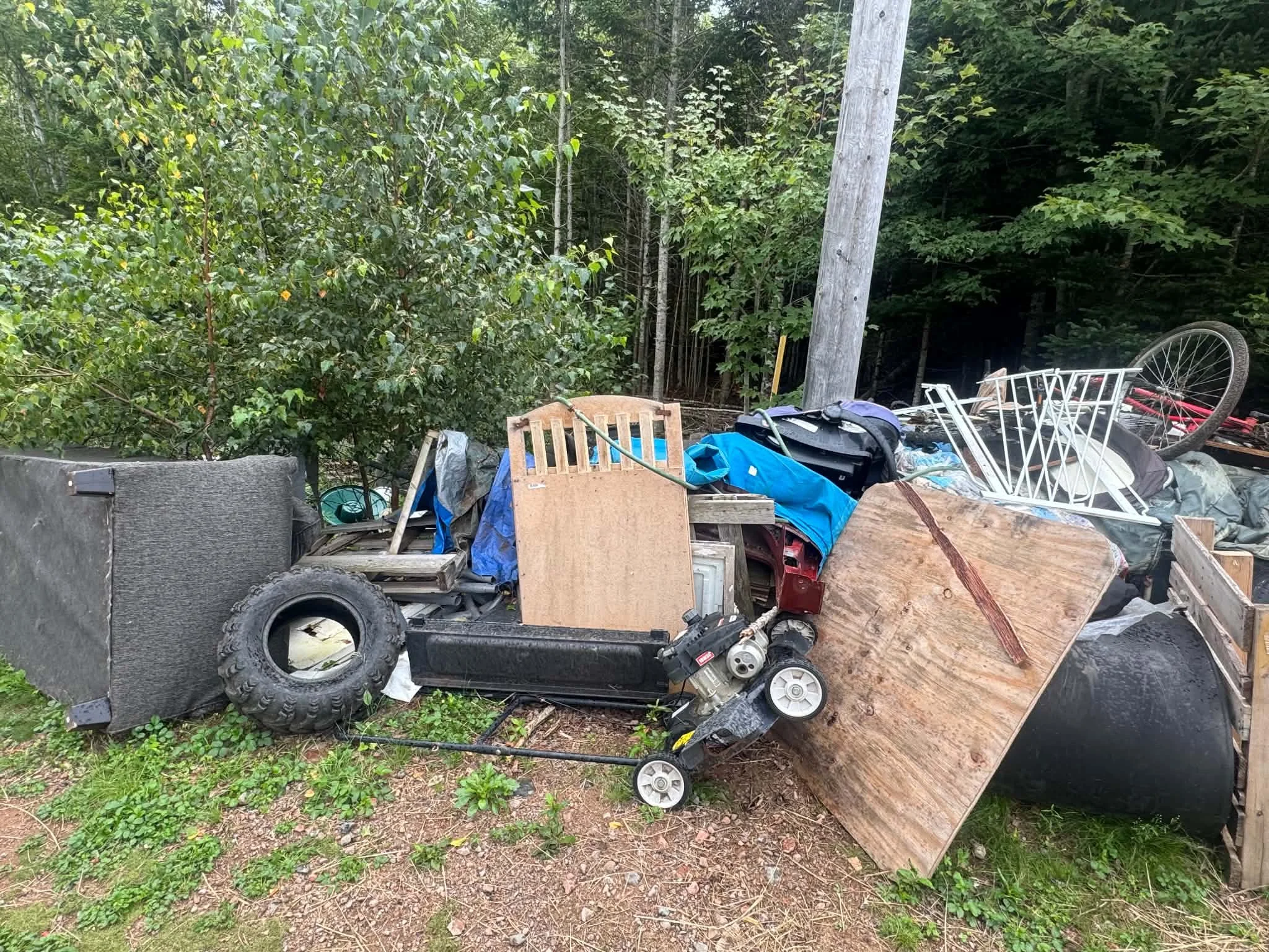 A pile of discarded items and debris outdoors next to a wooded area, including a bicycle wheel, an orange plastic chair, black speakers, a large plastic bag, pieces of wood, and various other miscellaneous objects.