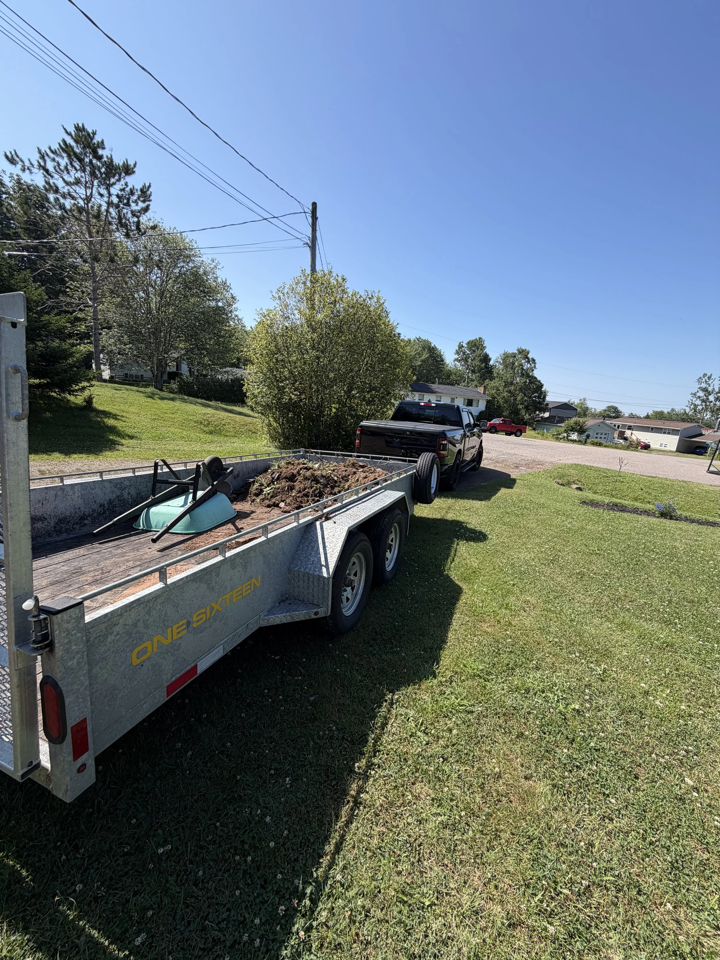 A trailer filled with dirt and gardening tools, attached to a black pickup truck, parked on a grassy area near residential houses and trees.