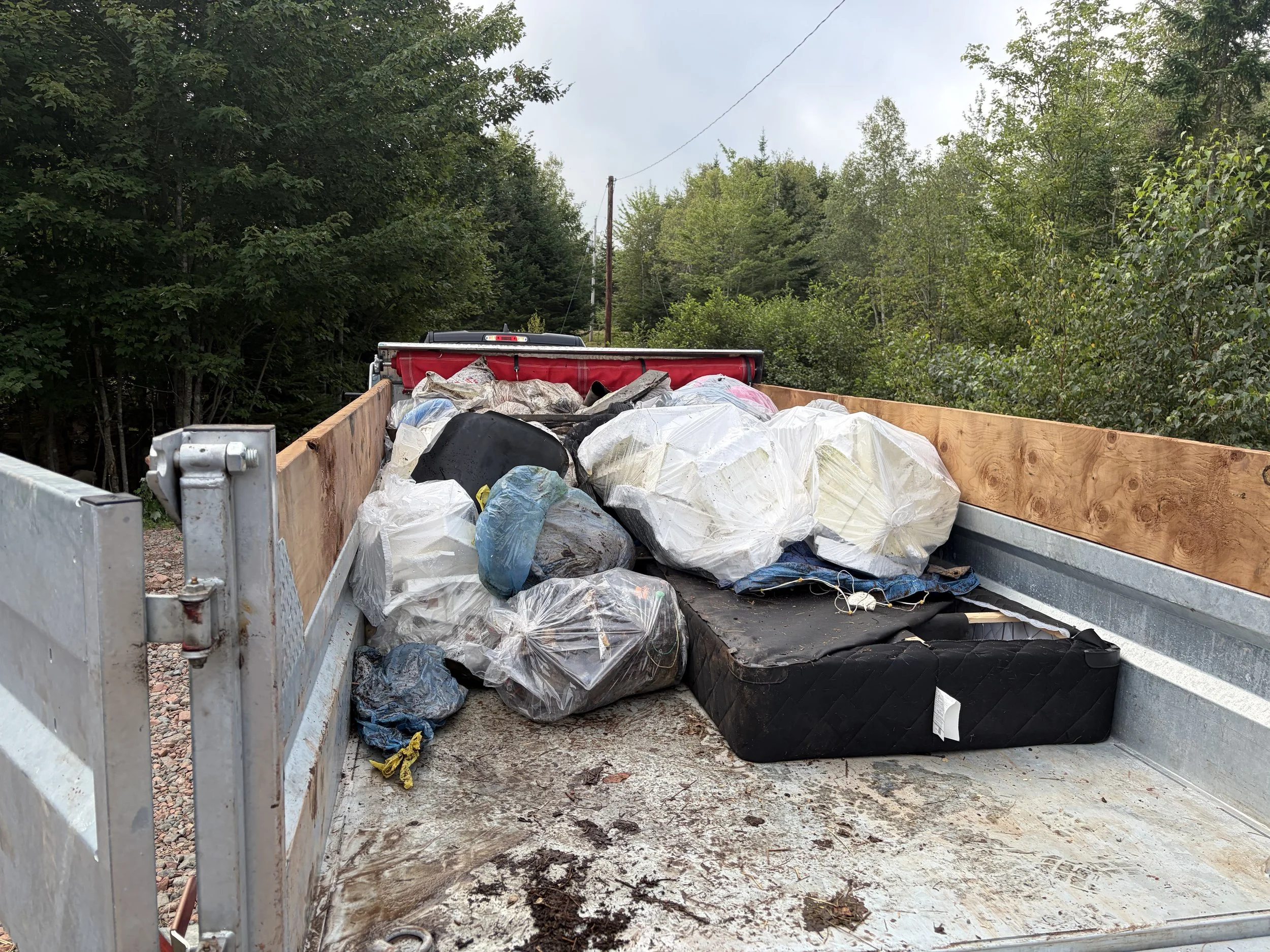 Flatbed trailer filled with trash bags, some black and some transparent, containing garbage, with a wooded area in the background.
