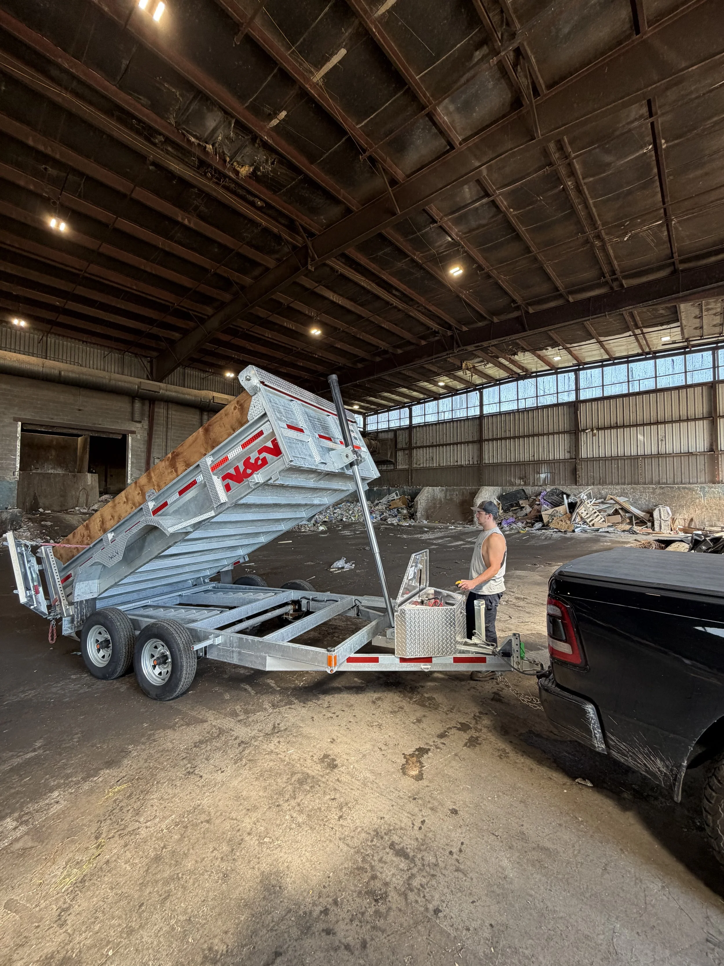 Inside a large industrial warehouse with a high ceiling, a man is operating a trailer with a hydraulic lift, lifting a large wooden beam. The warehouse has large windows near the ceiling letting in natural light, and there is a pile of debris and tra