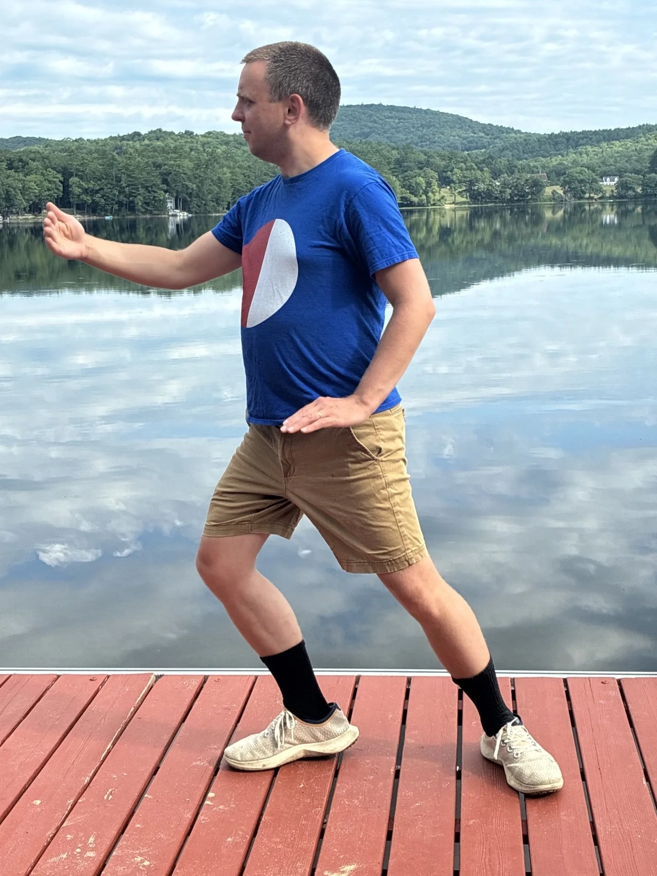 A man stands on a wooden dock by a lake, stretching his arm forward with a serious expression. He is wearing a blue t-shirt, tan shorts, black socks, and sneakers, with a backdrop of green trees and hills reflected in the water.