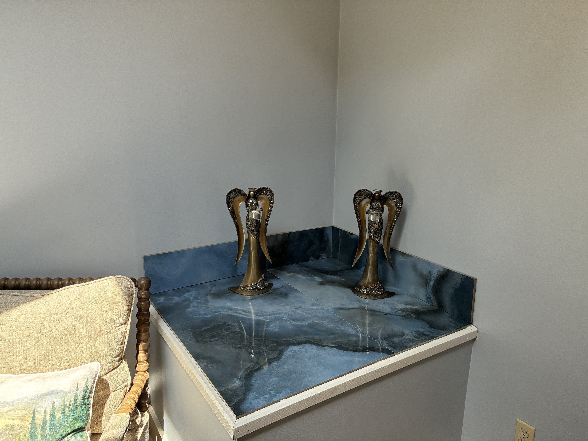 Decorative brass lamp bases with ornate designs placed on a dark marble surface, against a plain wall. Part of a beige sofa with a colorful cushion is visible in the foreground.